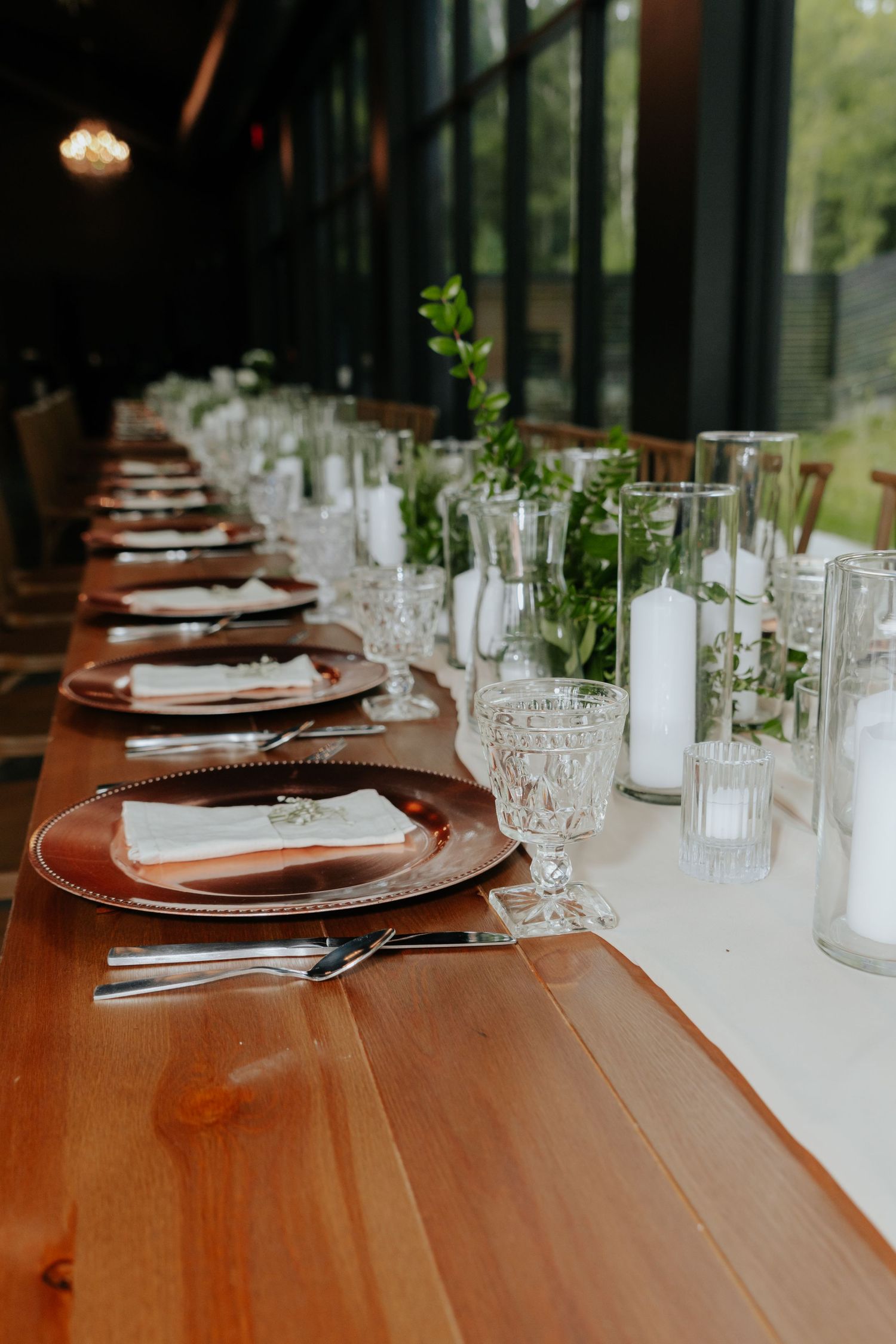Elegant wedding reception table with copper charger plates, crystal glassware, white candles, and greenery centerpieces.