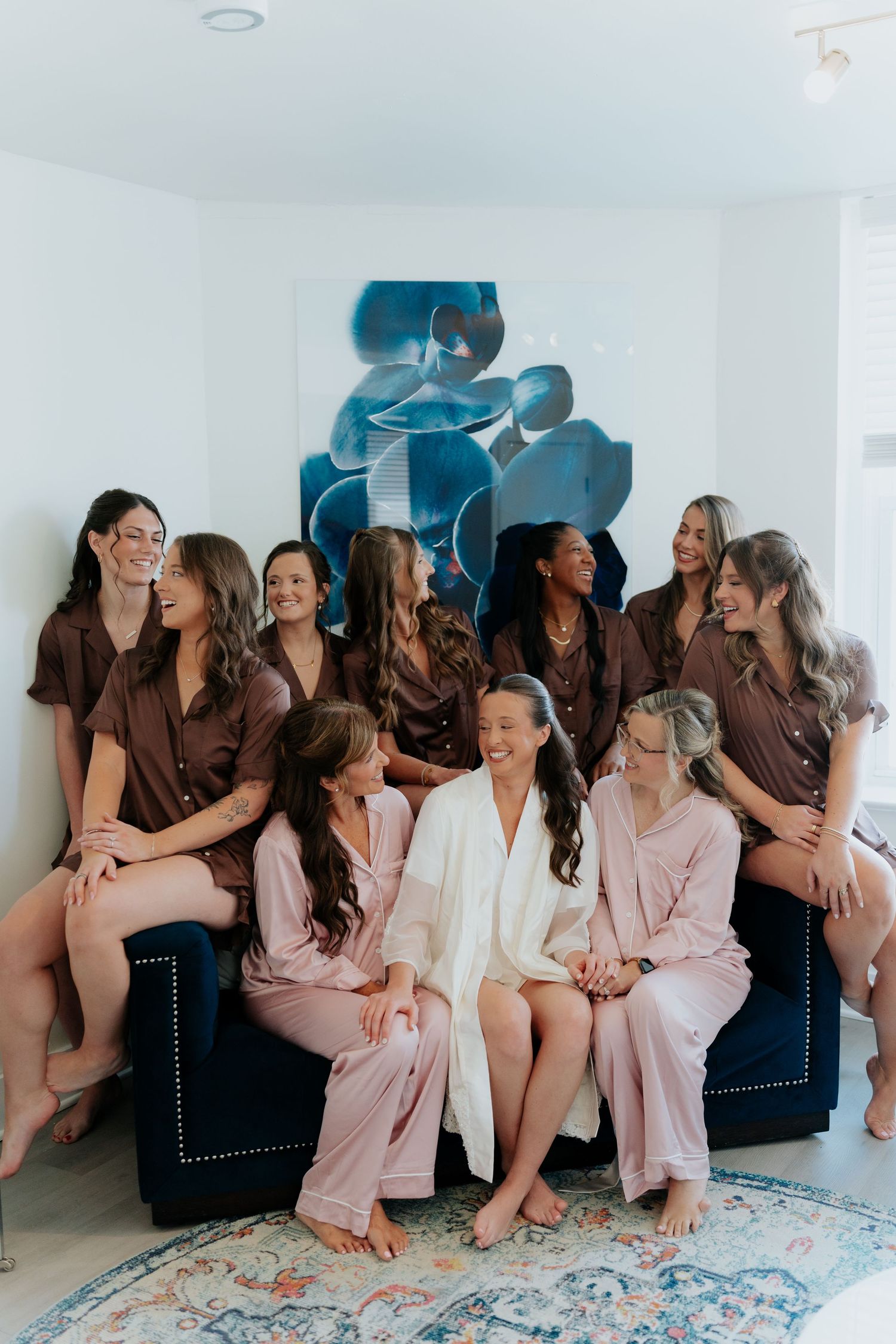Bride in white robe surrounded by bridesmaids in pink and brown satin pajamas laughing together before wedding.