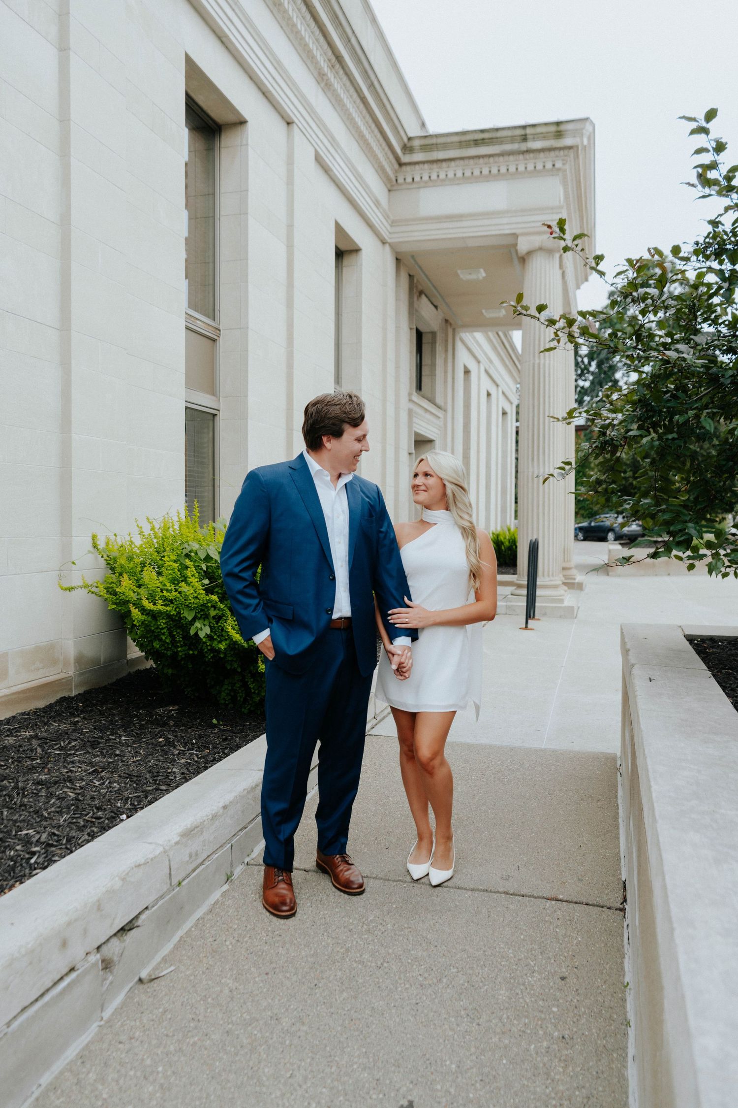 A couple shares a romantic moment in a white stone colonnade walkway during an engagement photoshoot.