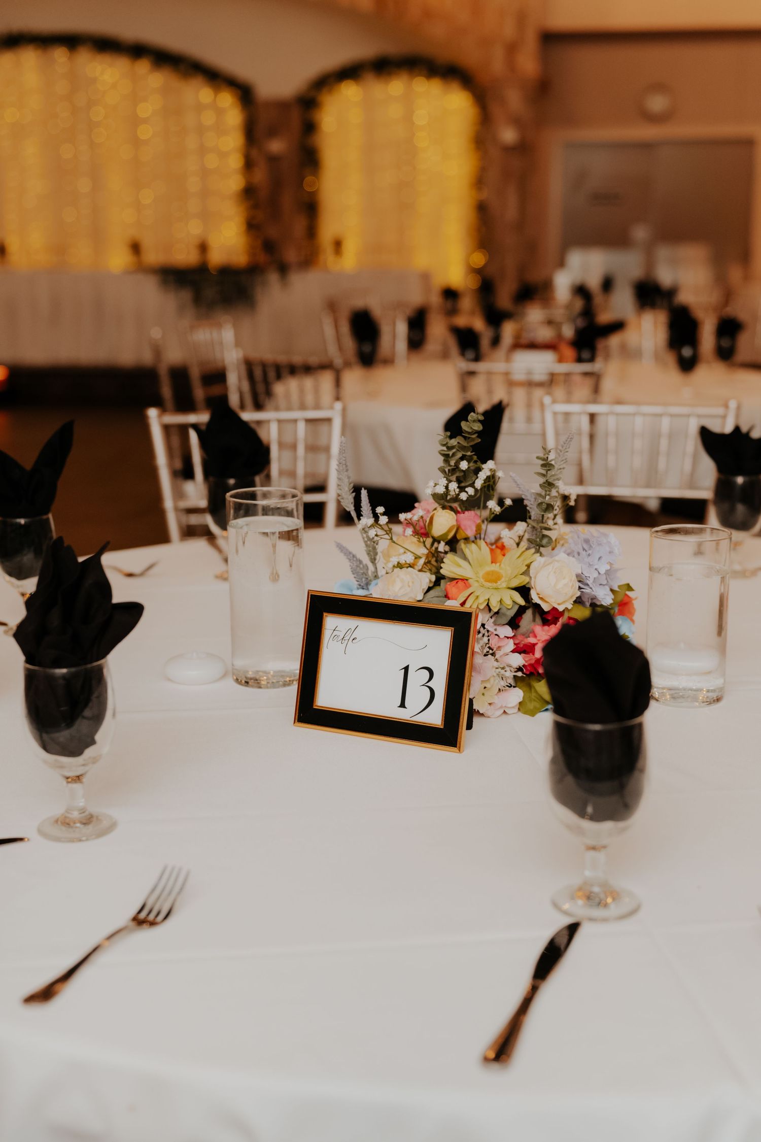 Elegant wedding reception table 13 with white linen, black napkins, floral centerpiece, and gold-trimmed place settings.