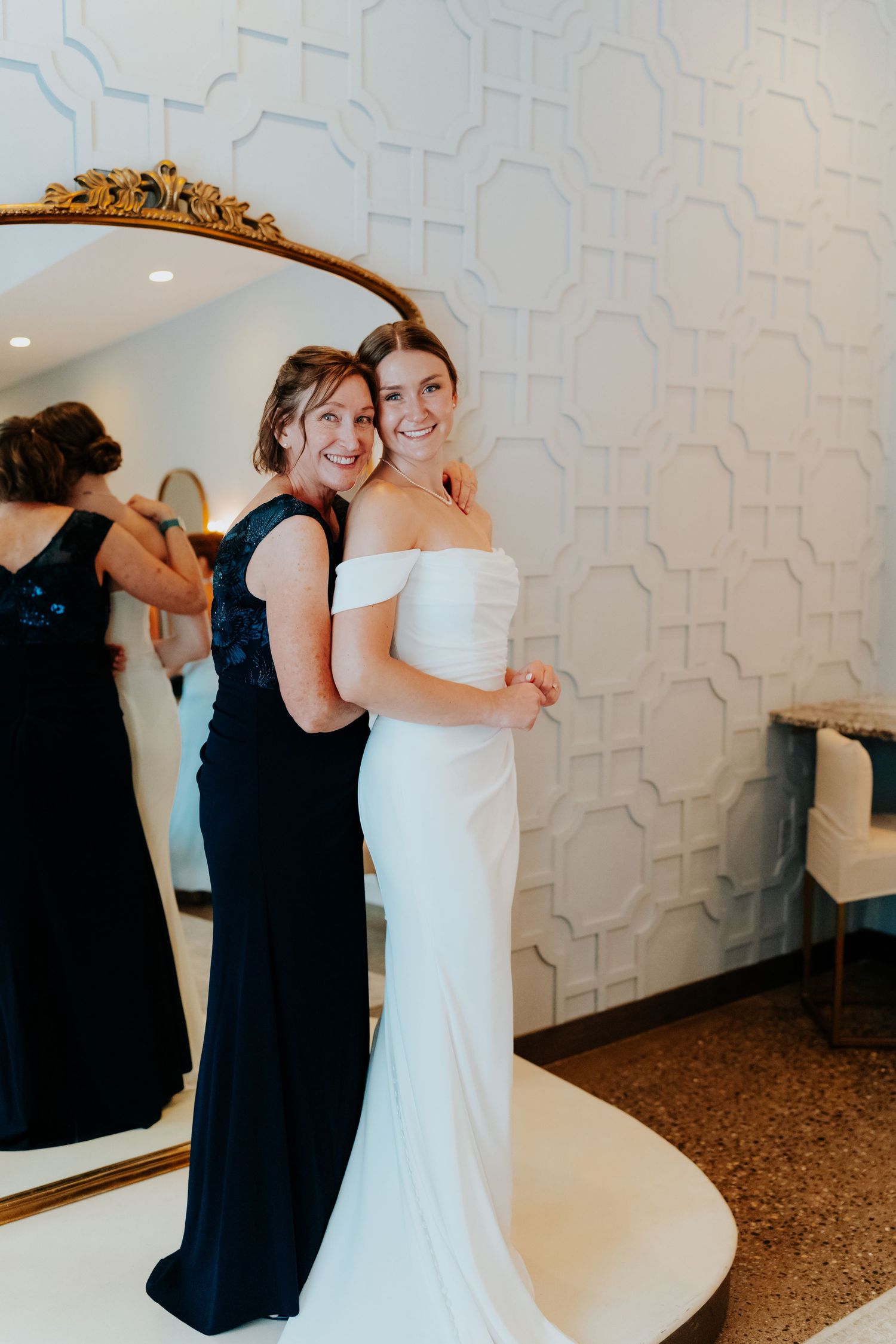 Bride in off-shoulder white gown smiles with woman in navy dress beside ornate gold mirror.