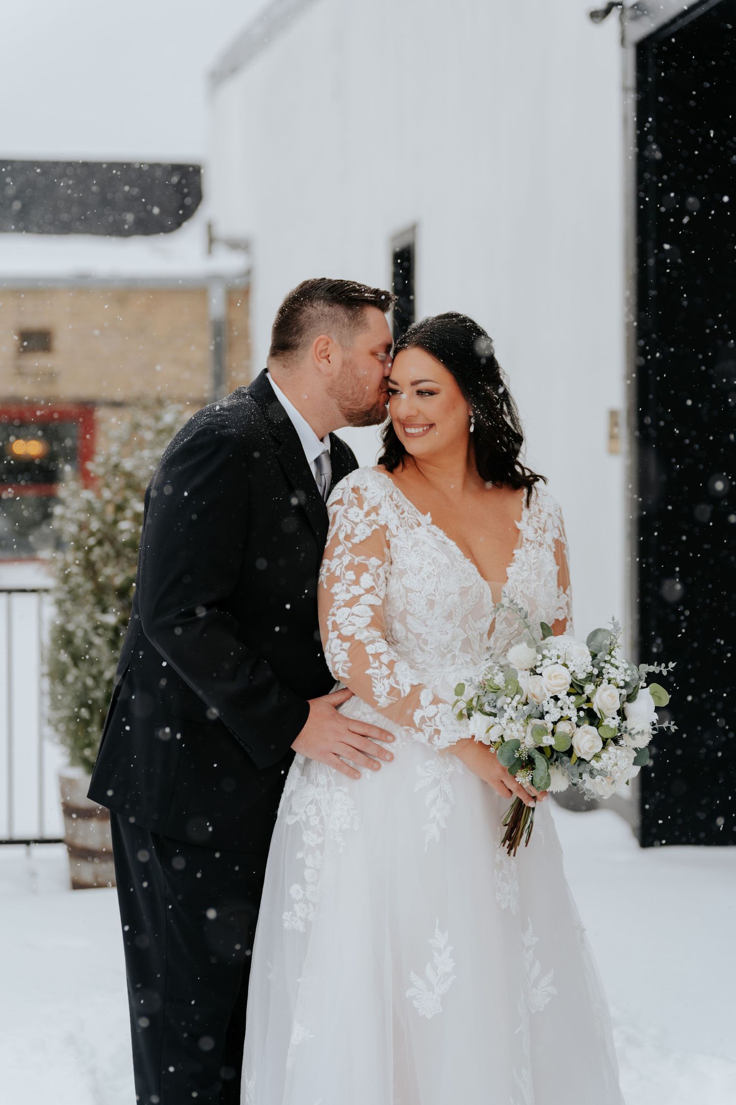 Bride and groom share a romantic kiss in the snow, bride wearing a lace long-sleeve wedding gown holding a white bouquet.