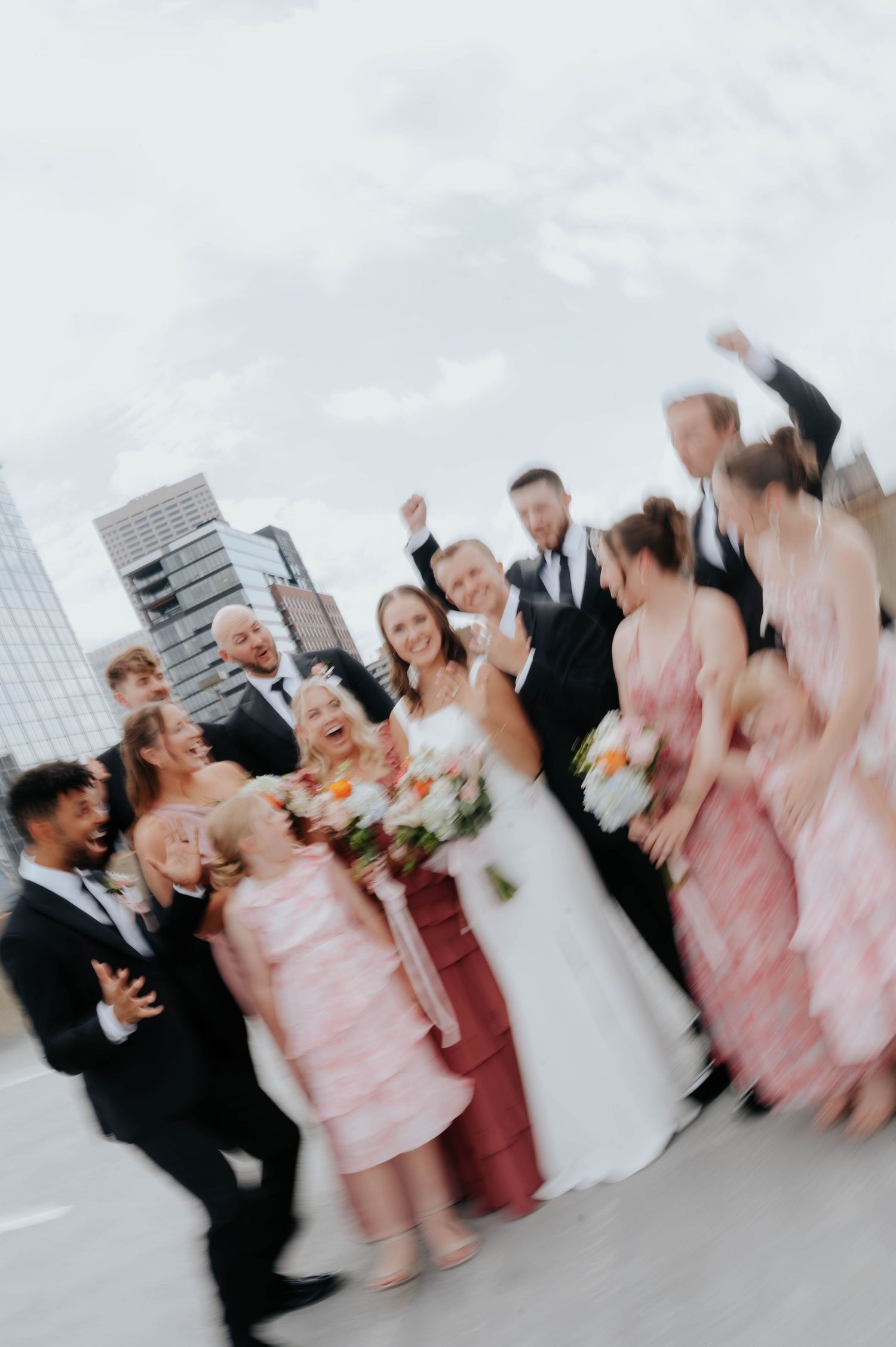 Joyful wedding party celebrating outdoors with bride in white gown, bridesmaids in pink dresses, and groomsmen in suits.