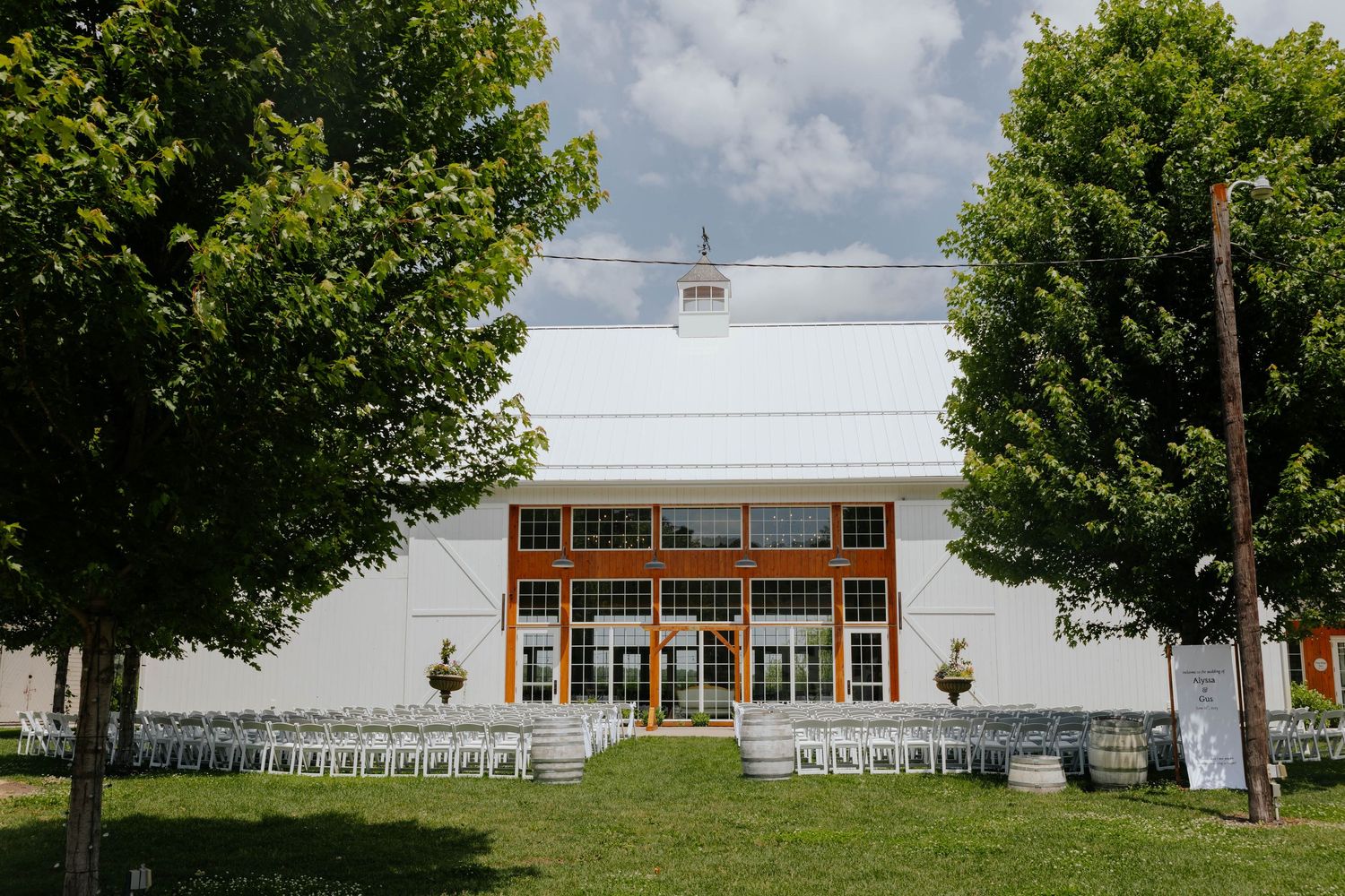 White barn wedding venue with orange-trimmed windows, rows of chairs on green lawn, framed by lush trees under cloudy sky.