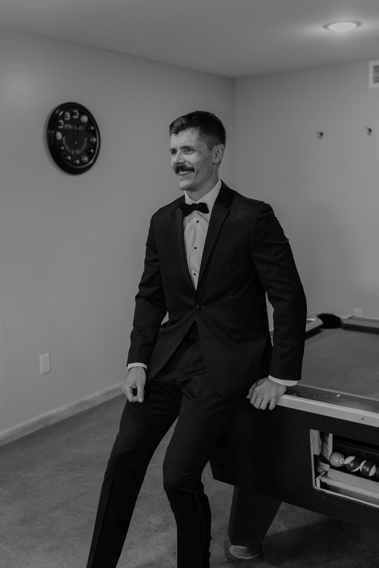 Groom in black tuxedo with bow tie smiling in a game room with pool table.