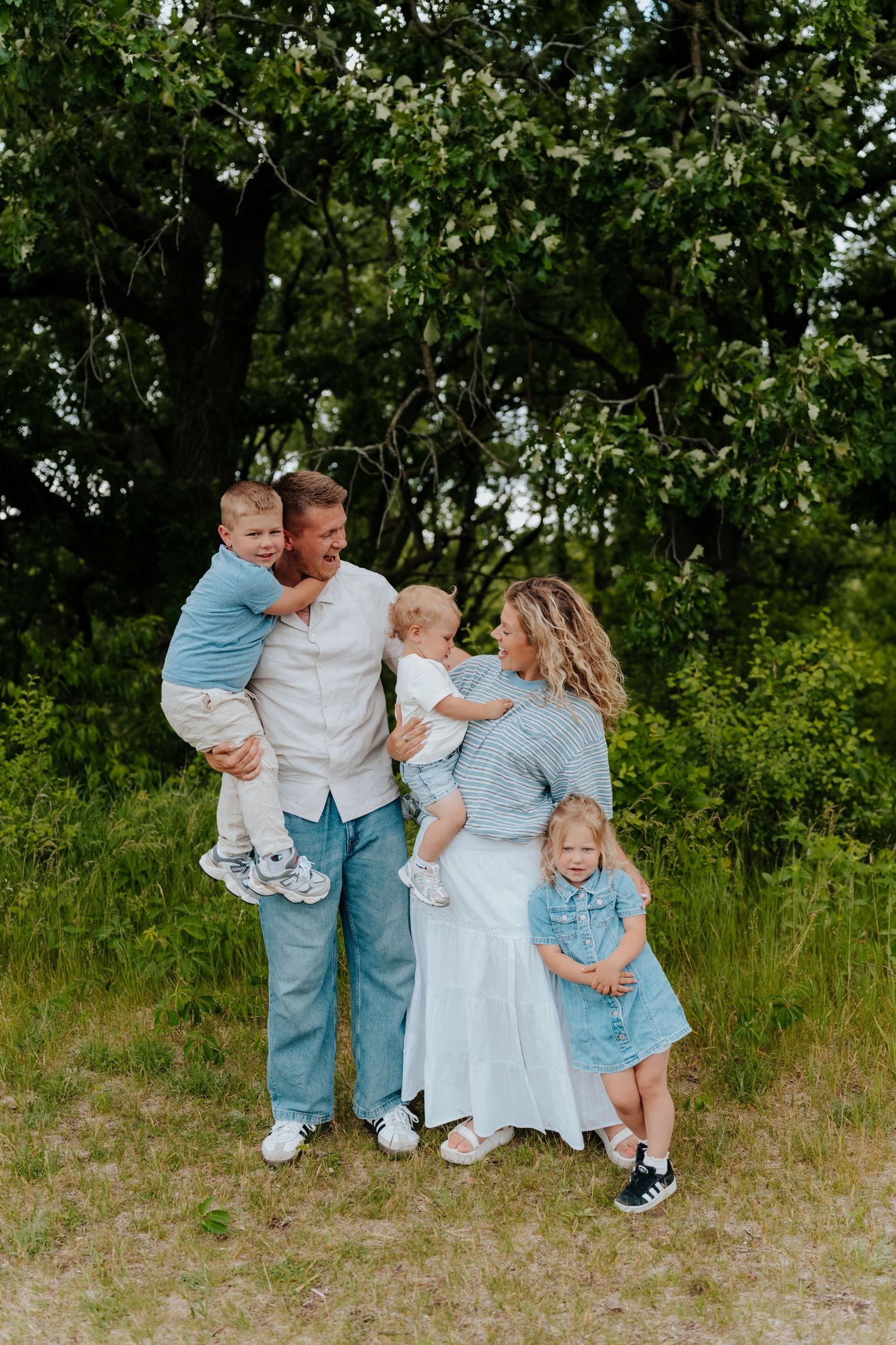 Happy family of five laughing outdoors, parents holding young children in a lush green park setting.