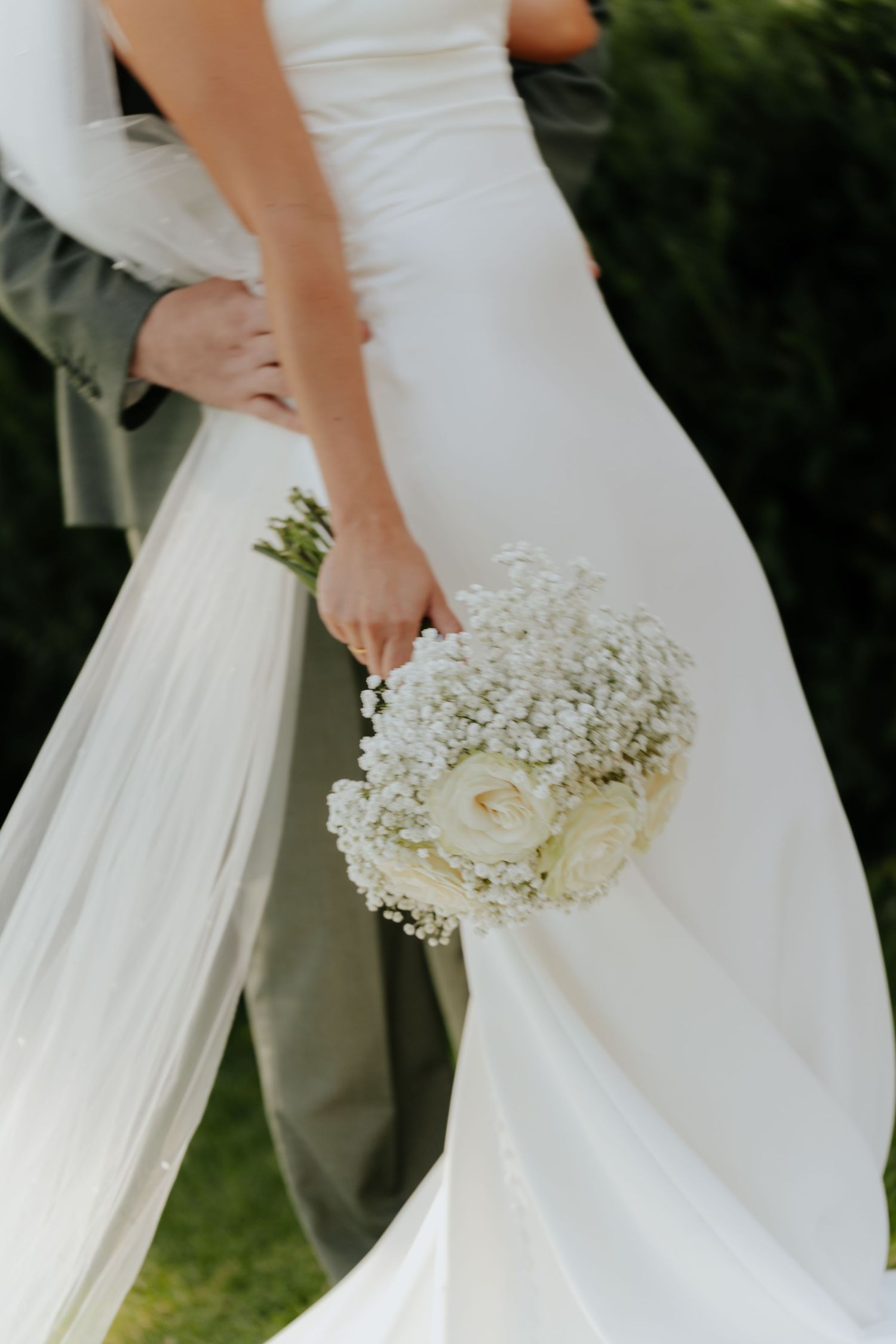 Bride in white gown holding a bouquet of white roses and baby's breath while groom embraces her waist outdoors.