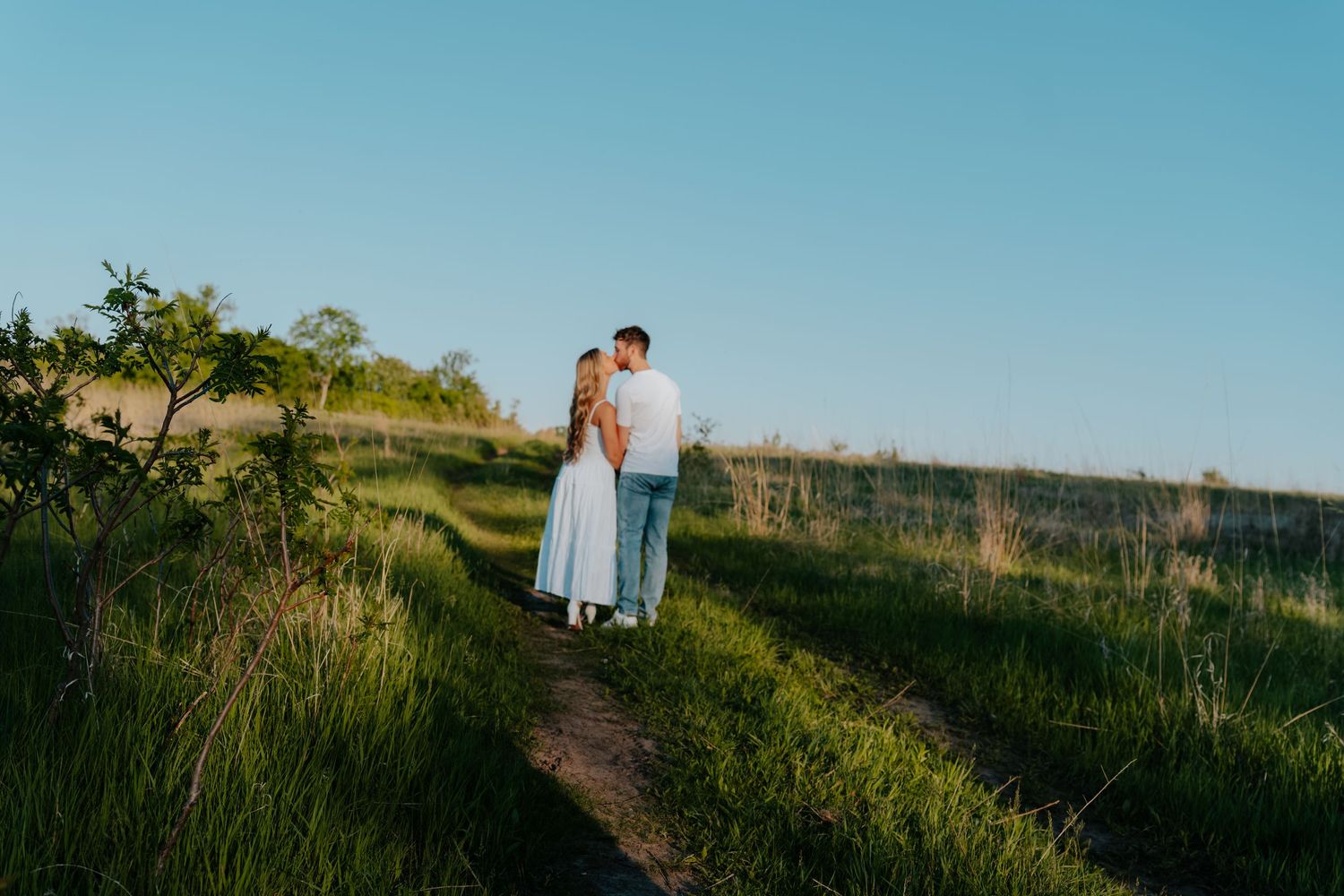 Couple embracing on a dirt path surrounded by lush green fields under a clear blue sky at golden hour.