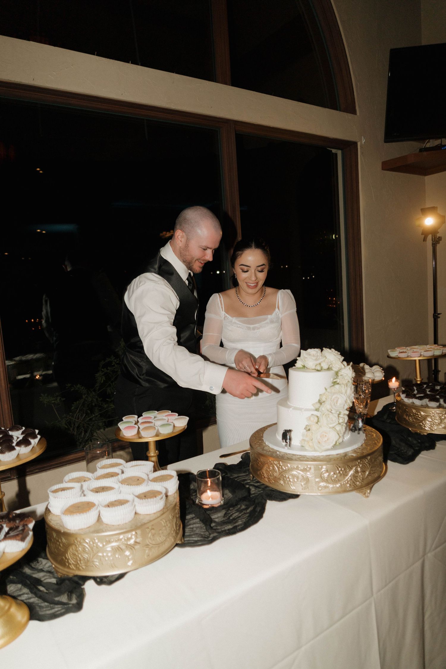 A couple in wedding attire serves cake to guests at a reception with multiple dessert plates on a long white table.