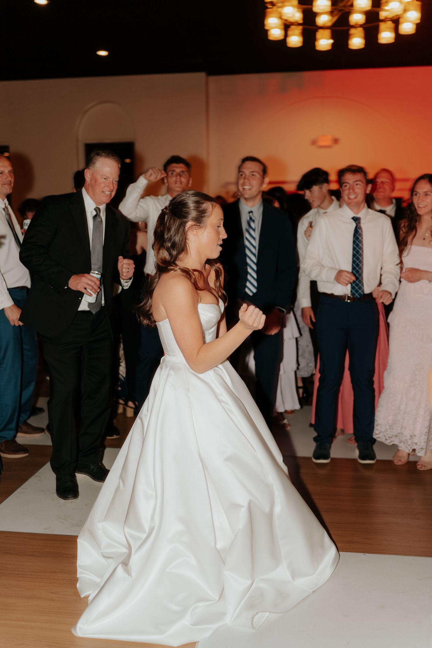 Bride in white ball gown dancing joyfully on the dance floor surrounded by wedding guests at reception.