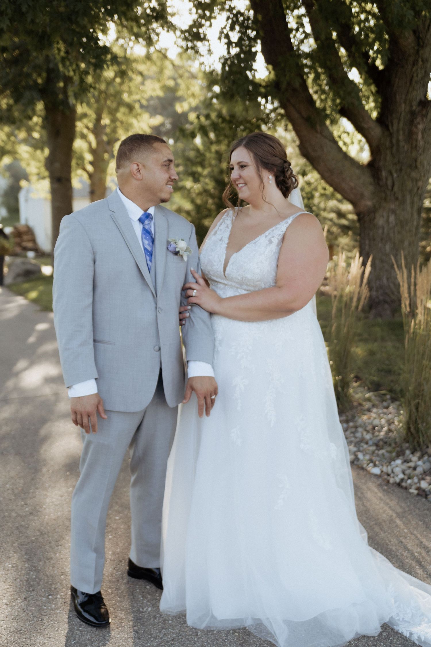 A couple shares a romantic moment on a tree-lined path, with the bride wearing a flowing white gown and groom in light gray suit.