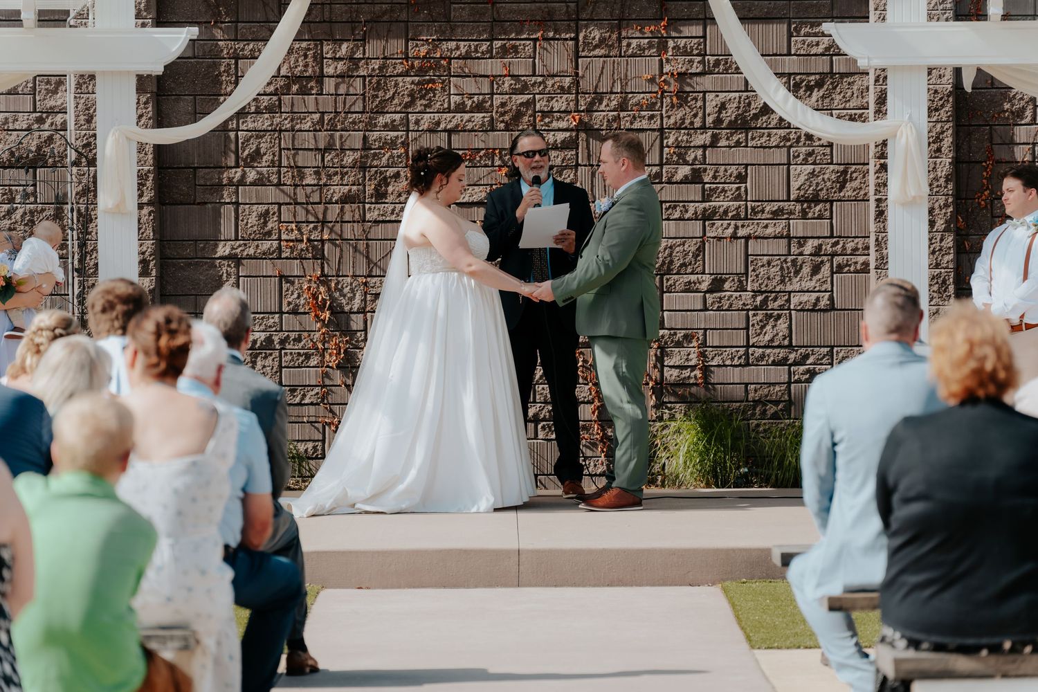 Outdoor wedding ceremony with bride and groom exchanging vows before officiant, guests seated on both sides, stone wall backdrop.