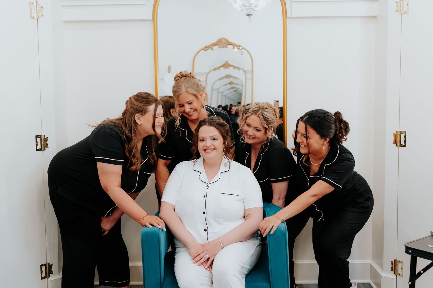 A bride in white sits smiling as four women in black uniforms lean in around her in an elegant salon.