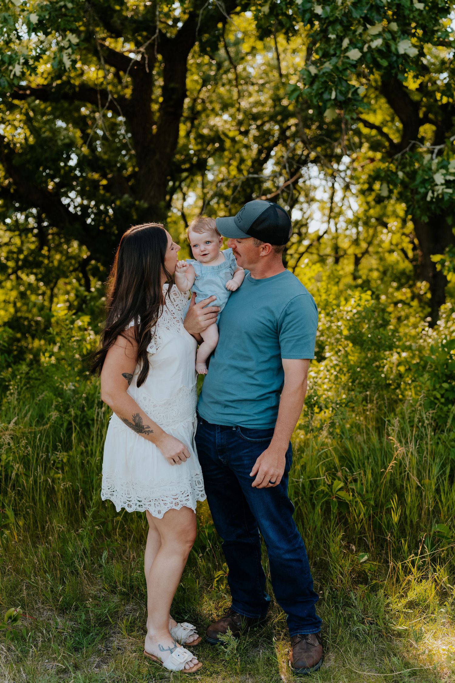 A young family poses outdoors in a lush green field, with parents holding their baby girl beneath sunlit trees.
