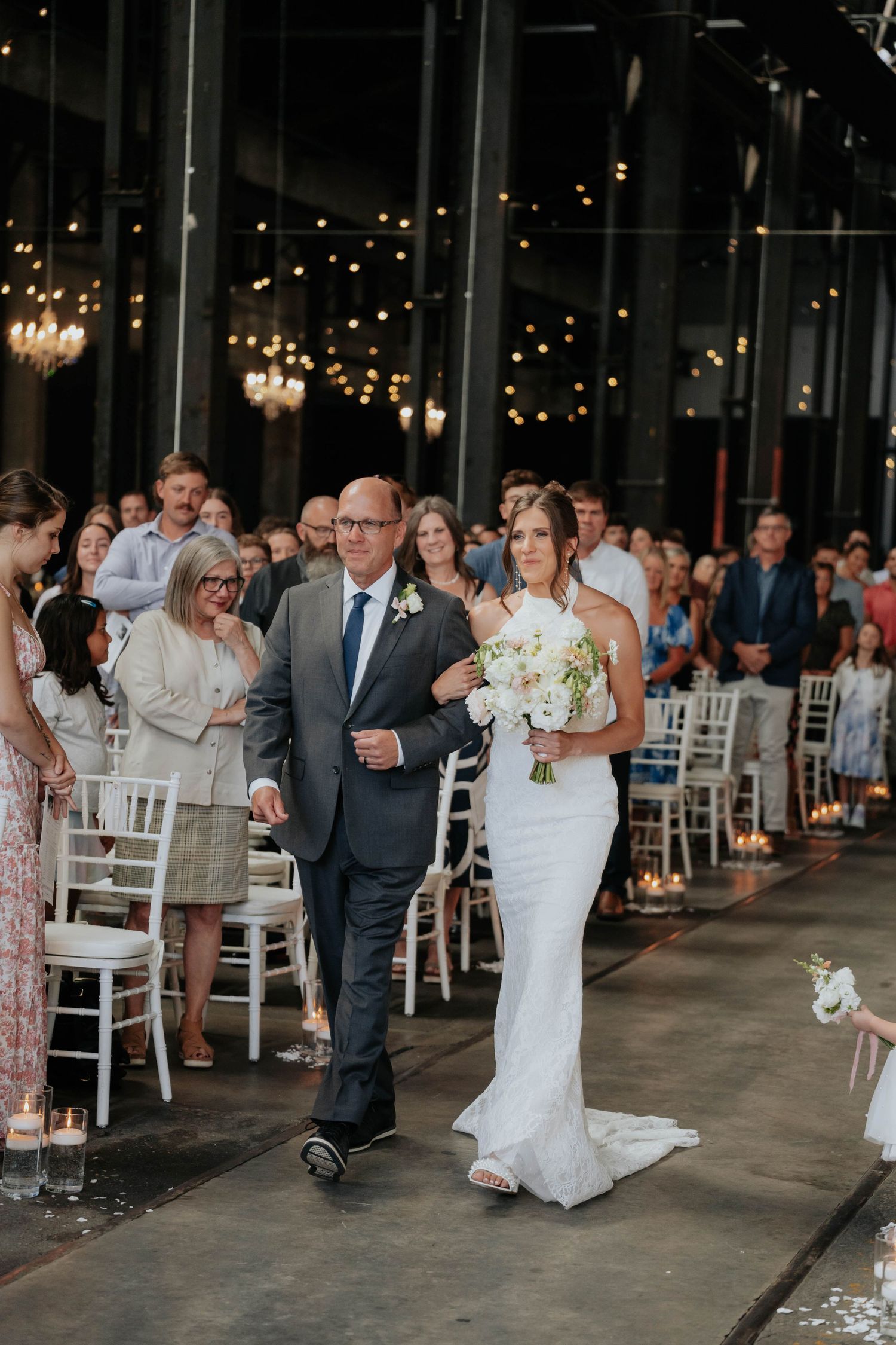 Bride walks down the aisle with her father in an elegant industrial wedding venue adorned with string lights.