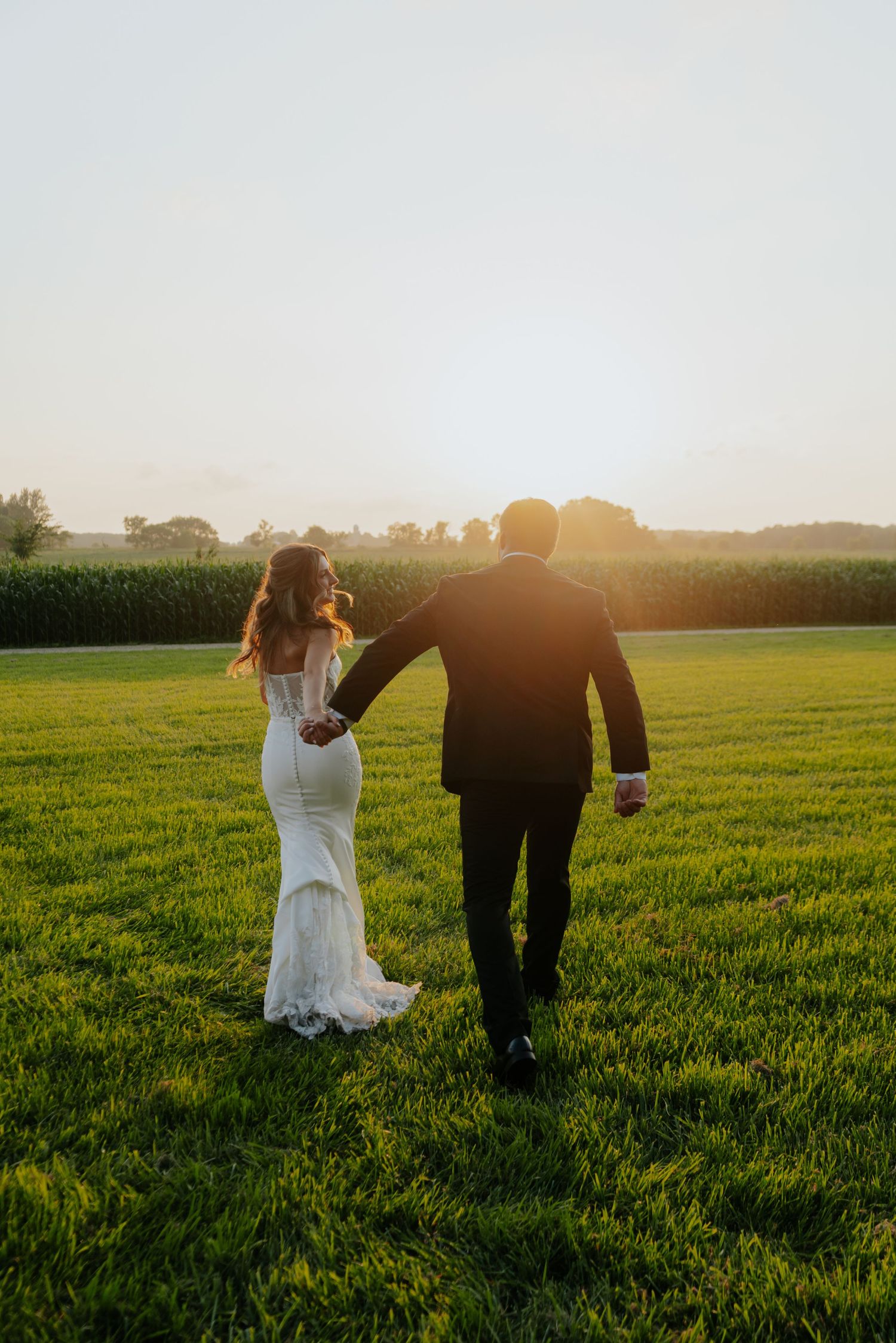 A bride and groom walk hand-in-hand through a lush green field at golden sunset, bride wearing elegant lace gown.