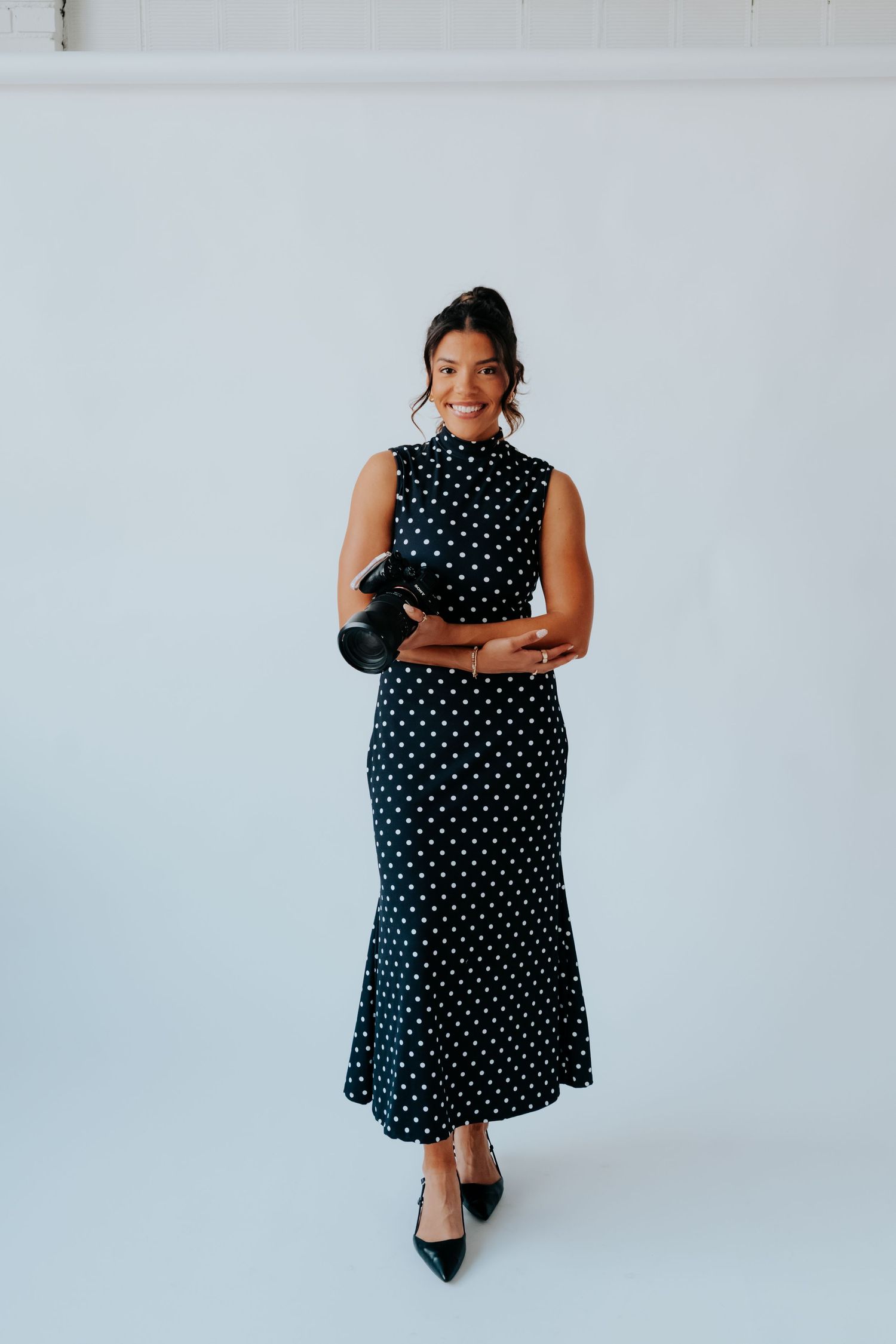 Woman wearing a black and white polka dot midi dress with heels, holding a camera in a studio setting.