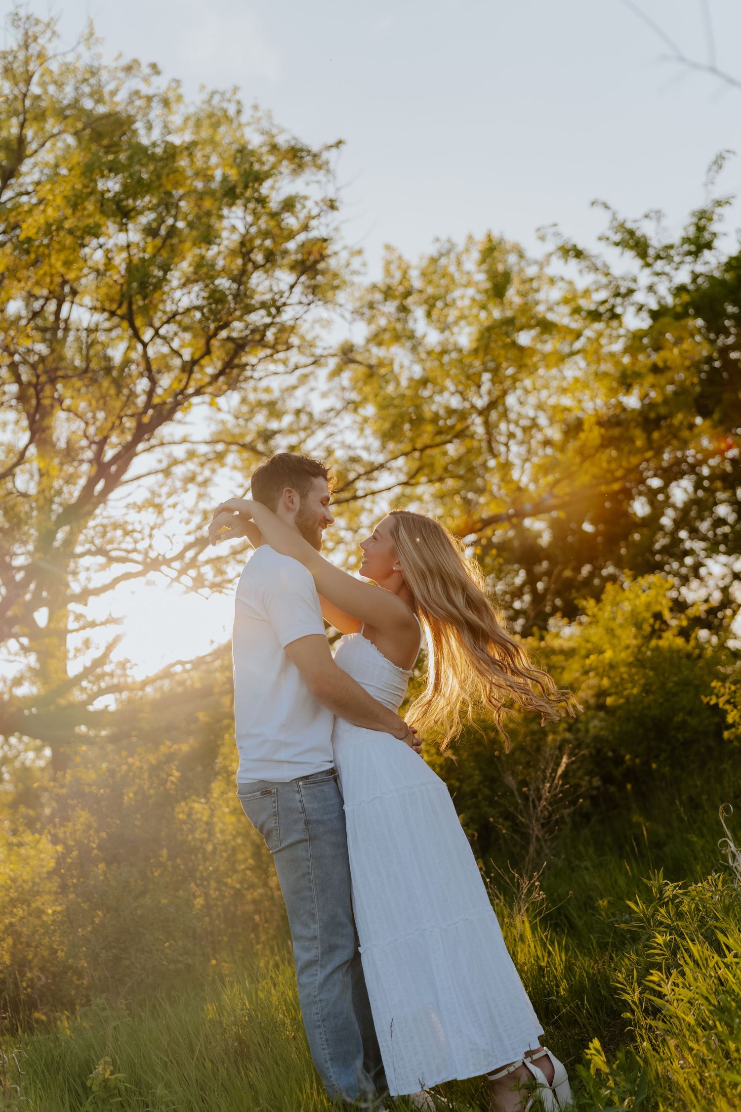 Couple embracing in golden sunset light surrounded by lush green trees, woman in white dress, man in white tee and jeans.