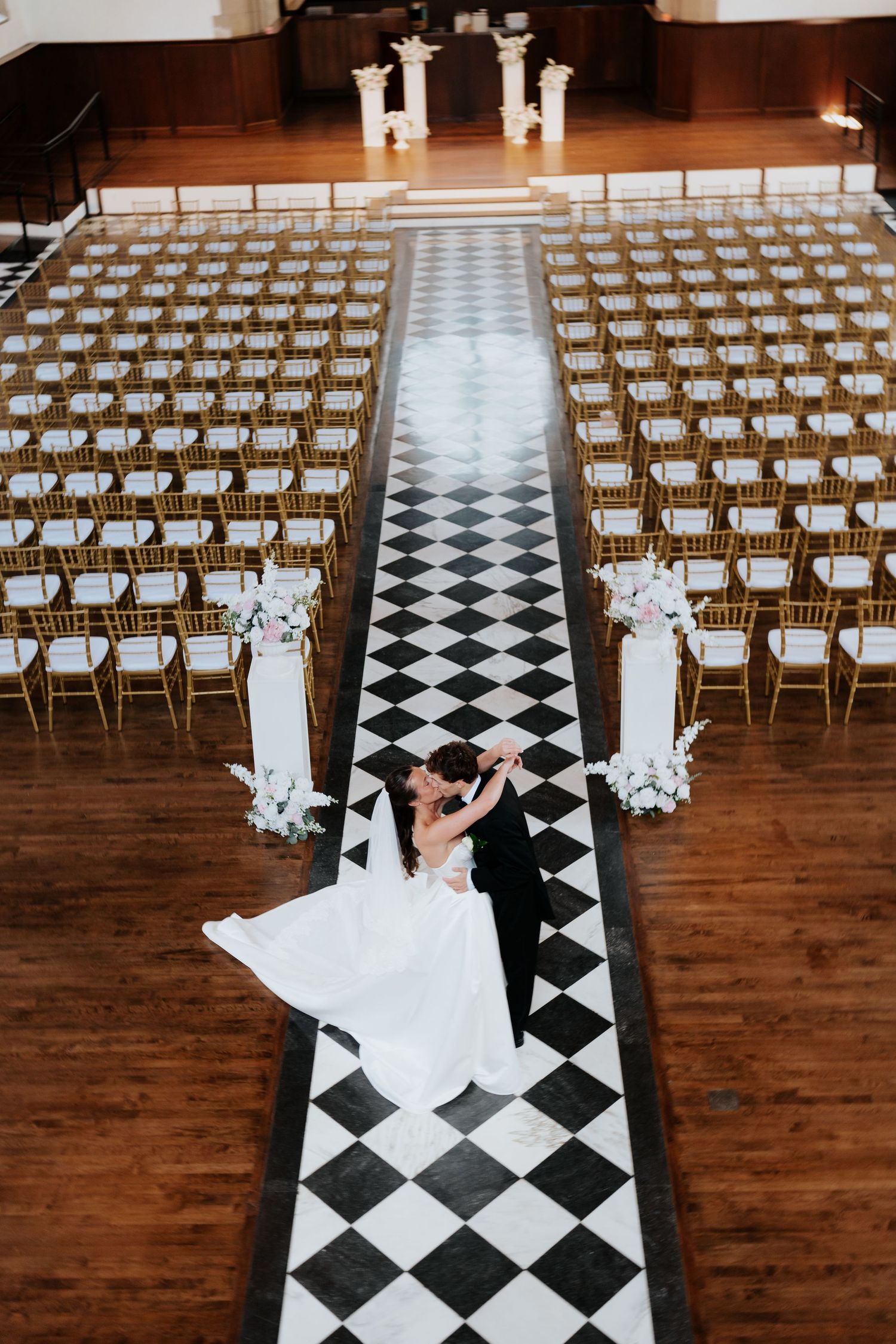 Bride and groom dancing on black and white checkered aisle in elegant wedding venue with rows of gold chairs.