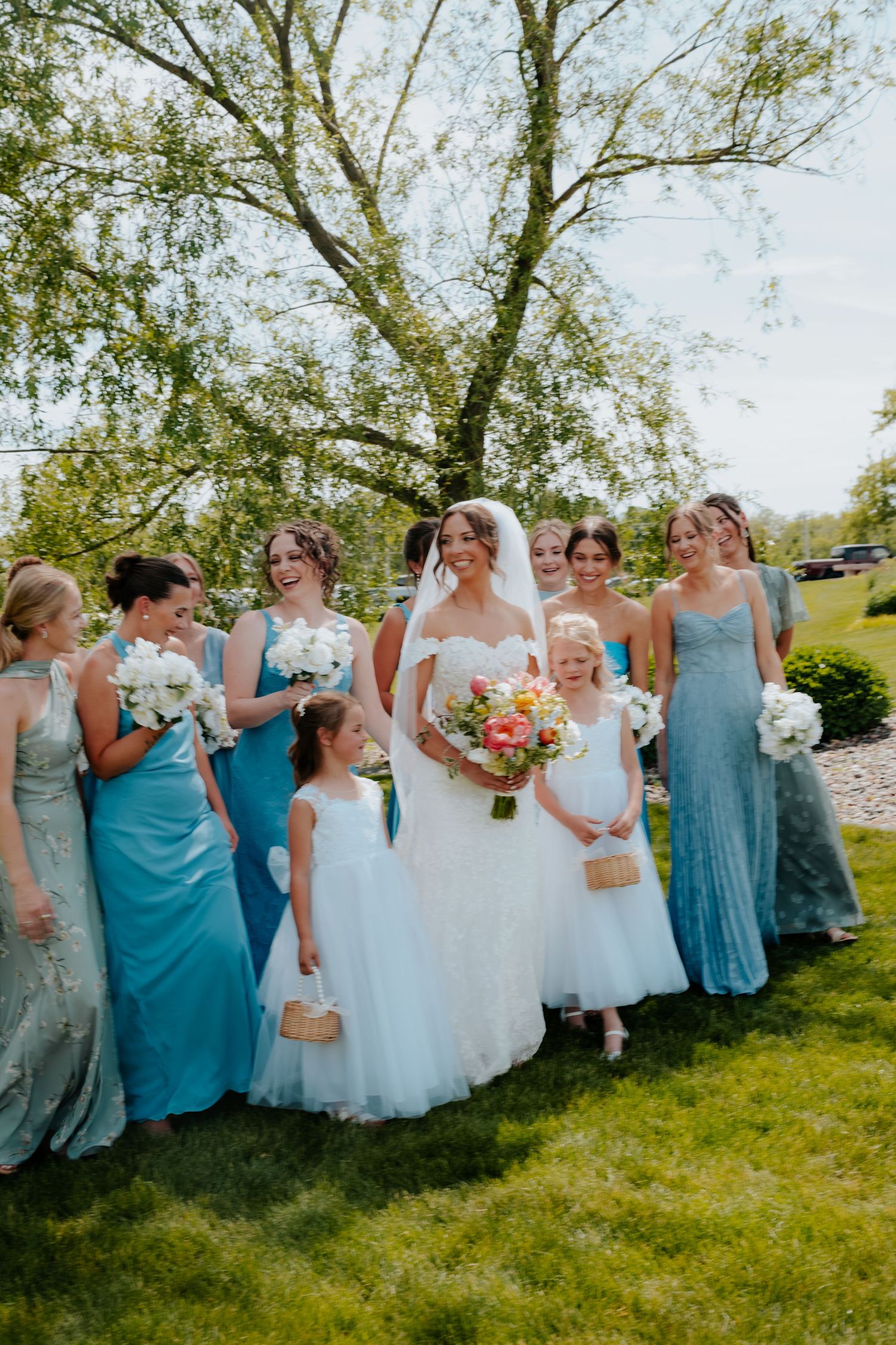 Bride in white gown with bridesmaids in blue dresses and two flower girls outdoors on a sunny wedding day.