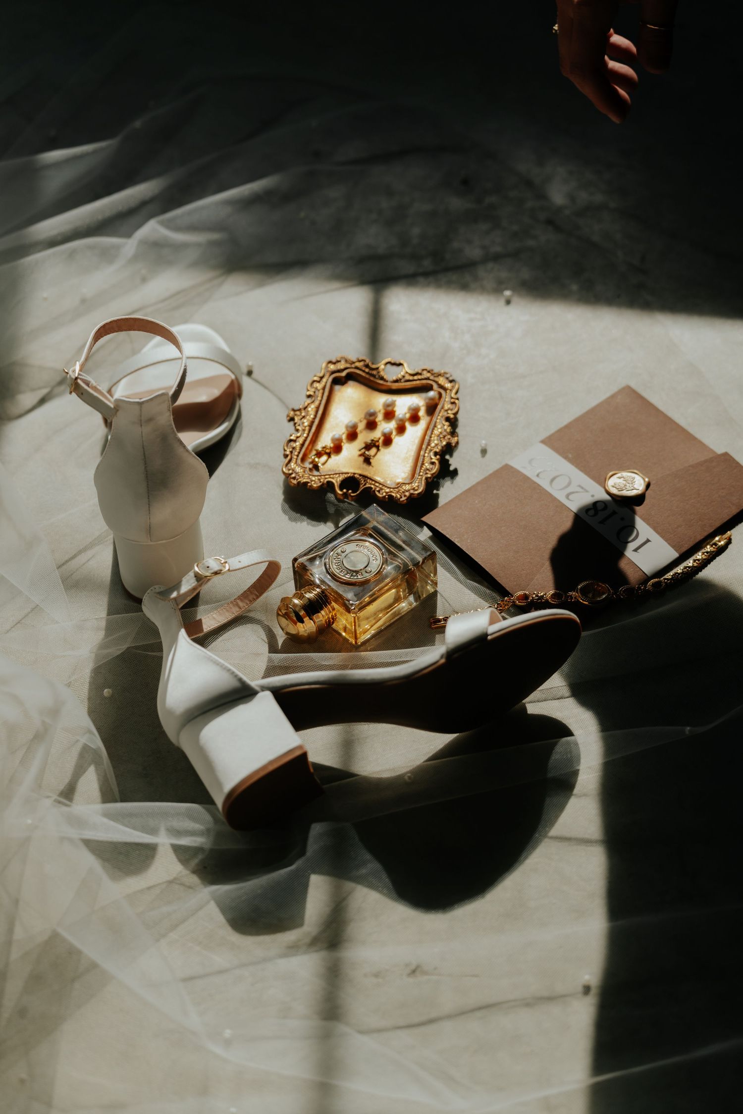 Bridal flat lay with white heels, gold perfume bottle, ornate jewelry box, and accessories on marble floor in soft light.