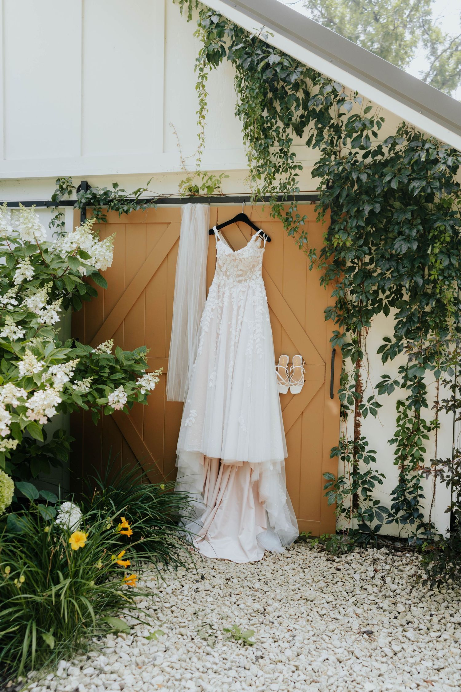 A lace wedding dress hangs on a barn door surrounded by lush greenery and white flowers with bridal heels nearby.