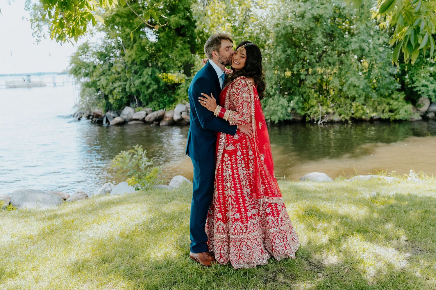 Couple sharing a romantic kiss outdoors near a river, bride in a stunning red Indian bridal lehenga, groom in a navy suit.