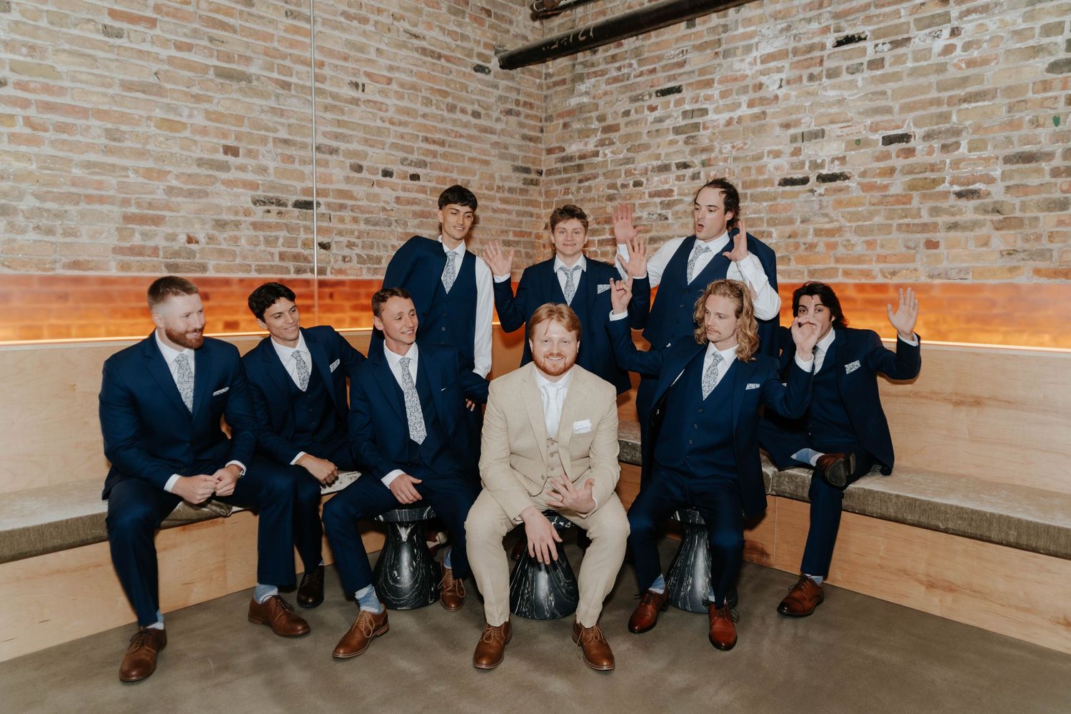 Groom in tan suit poses with groomsmen in navy suits on benches against exposed brick wall.