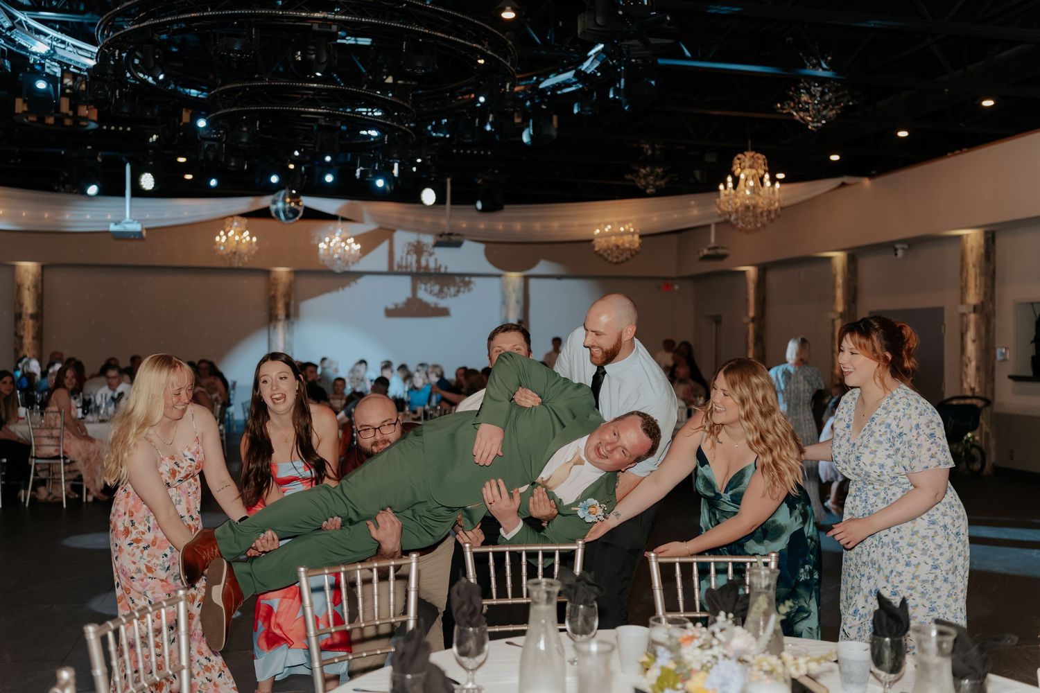 Wedding guests playfully lift a man in green suit at a elegant reception venue with chandeliers.