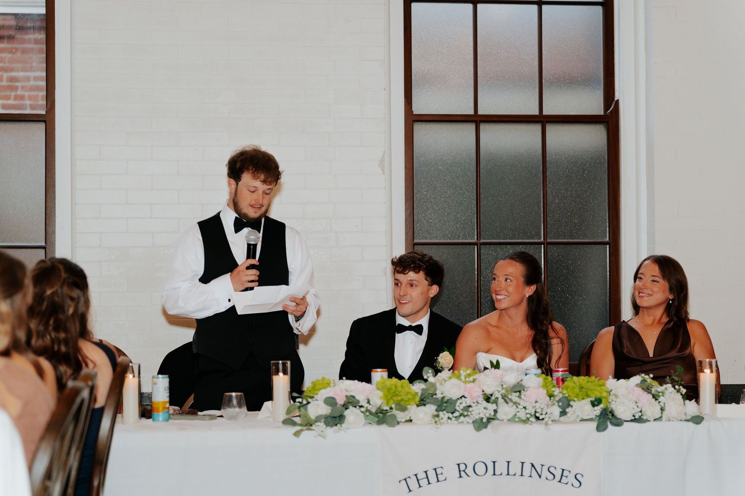 Best man giving a toast at The Rollinses wedding reception, with bride and groom seated at the head table.