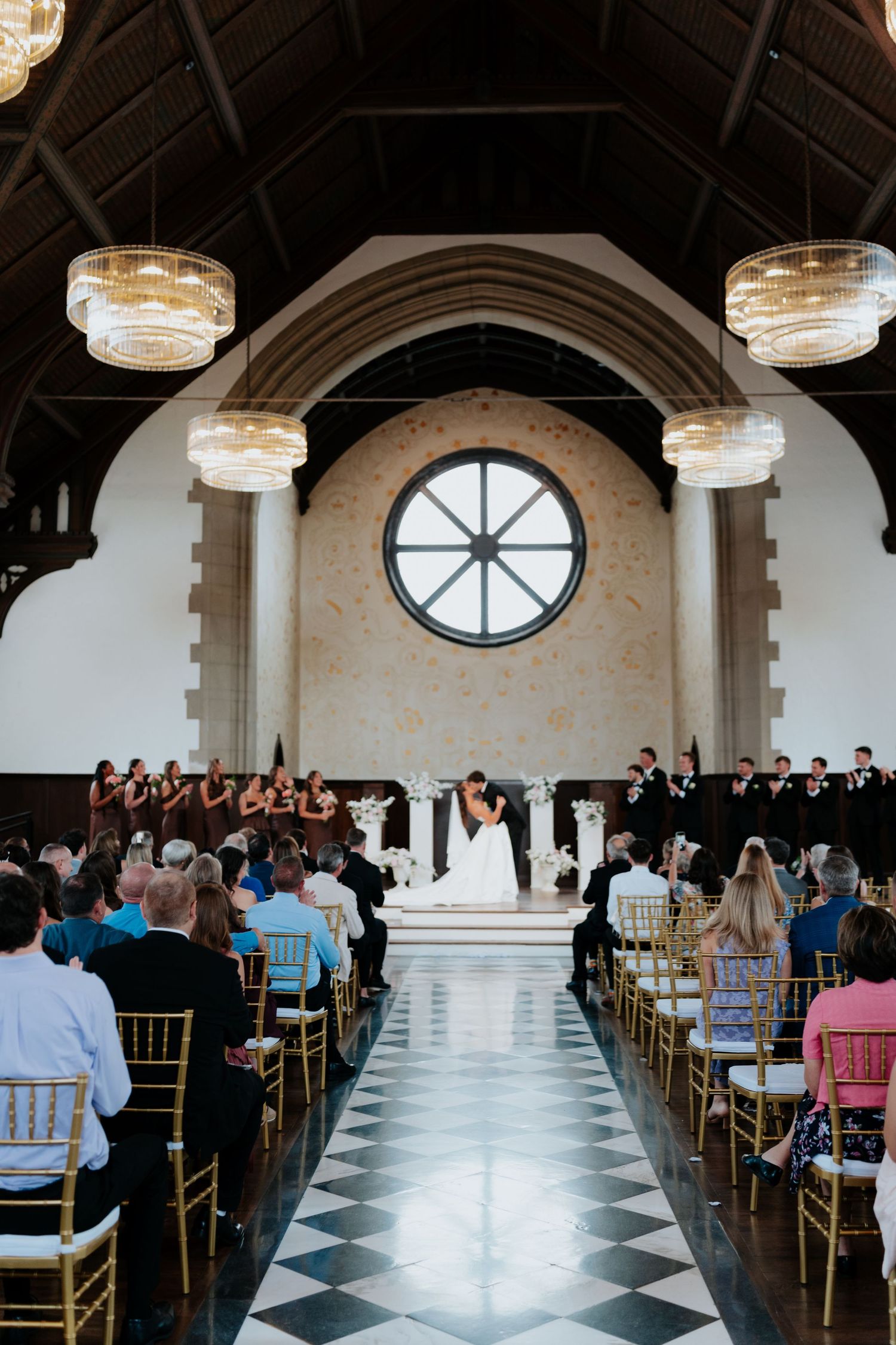 Elegant wedding ceremony in a grand hall with checkered floors, gold chairs, chandeliers, and couple at floral altar.