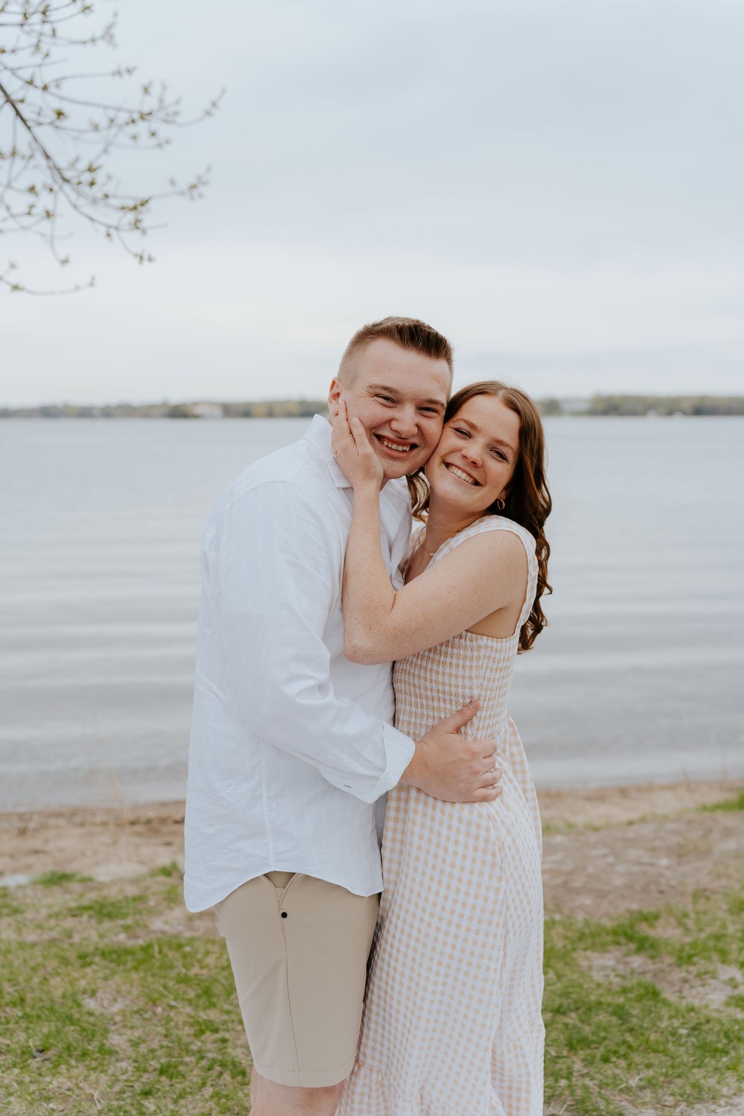 Happy couple embracing and smiling by a lakeside, woman in pink dress hugging man in white shirt outdoors.
