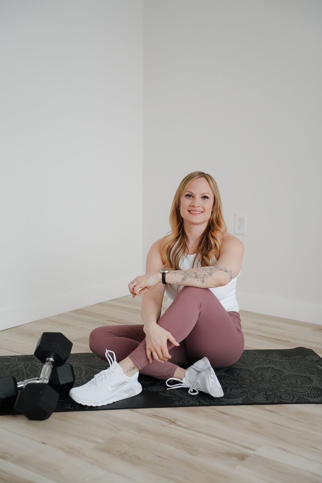 Woman in pink leggings and white top sits on yoga mat with dumbbells, smiling in bright home gym.