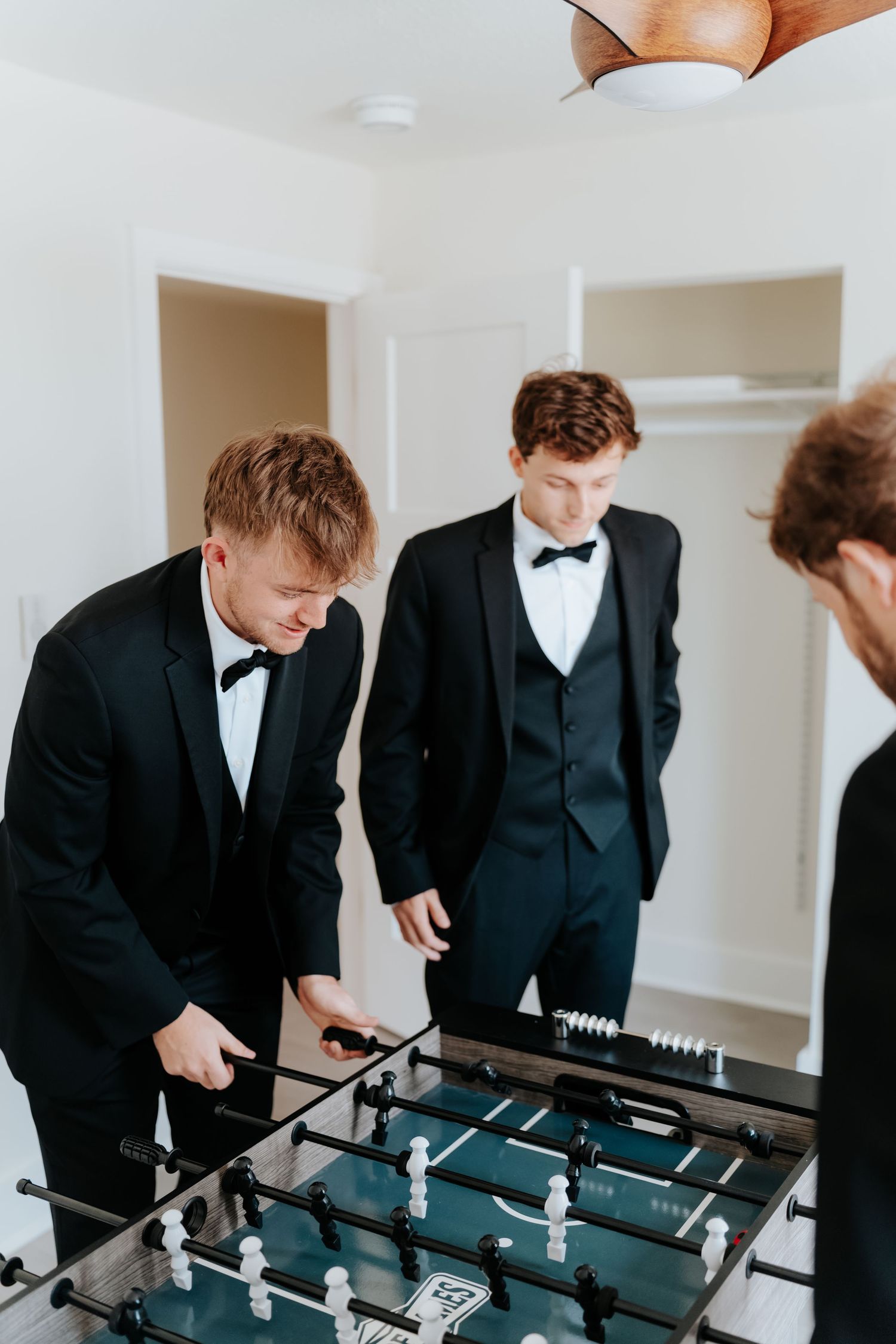 Three young men in black tuxedos playing foosball together before a formal event.
