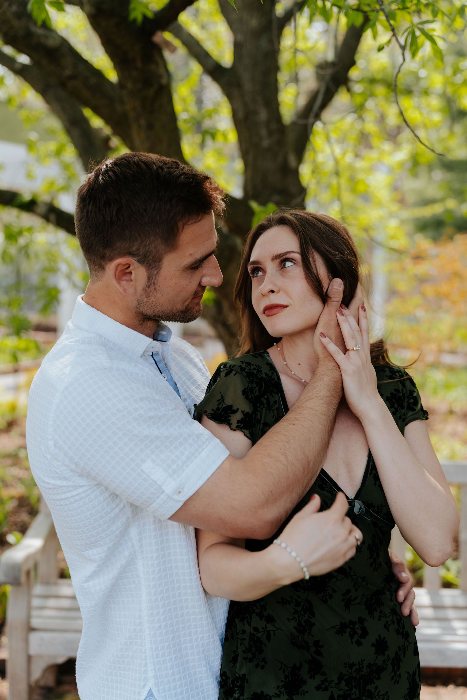 A couple shares a romantic moment outdoors, man gently holding woman's face near a tree.