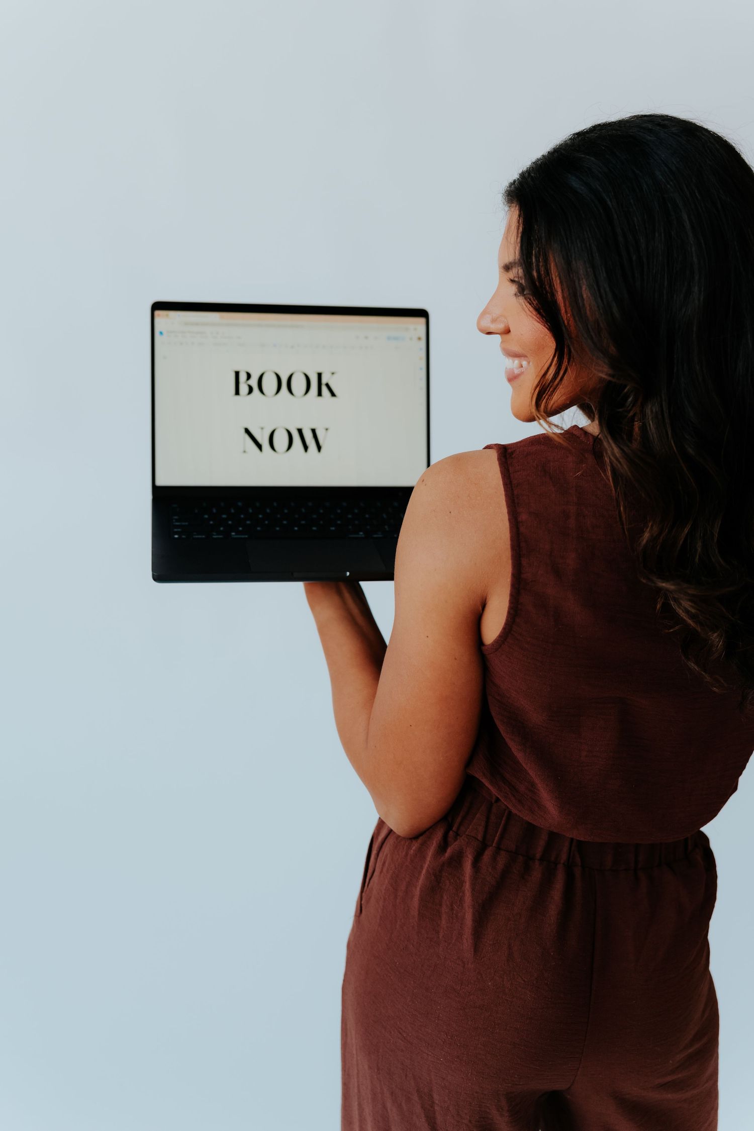 Woman in brown dress holding laptop displaying Book Now text, smiling over shoulder against light background.