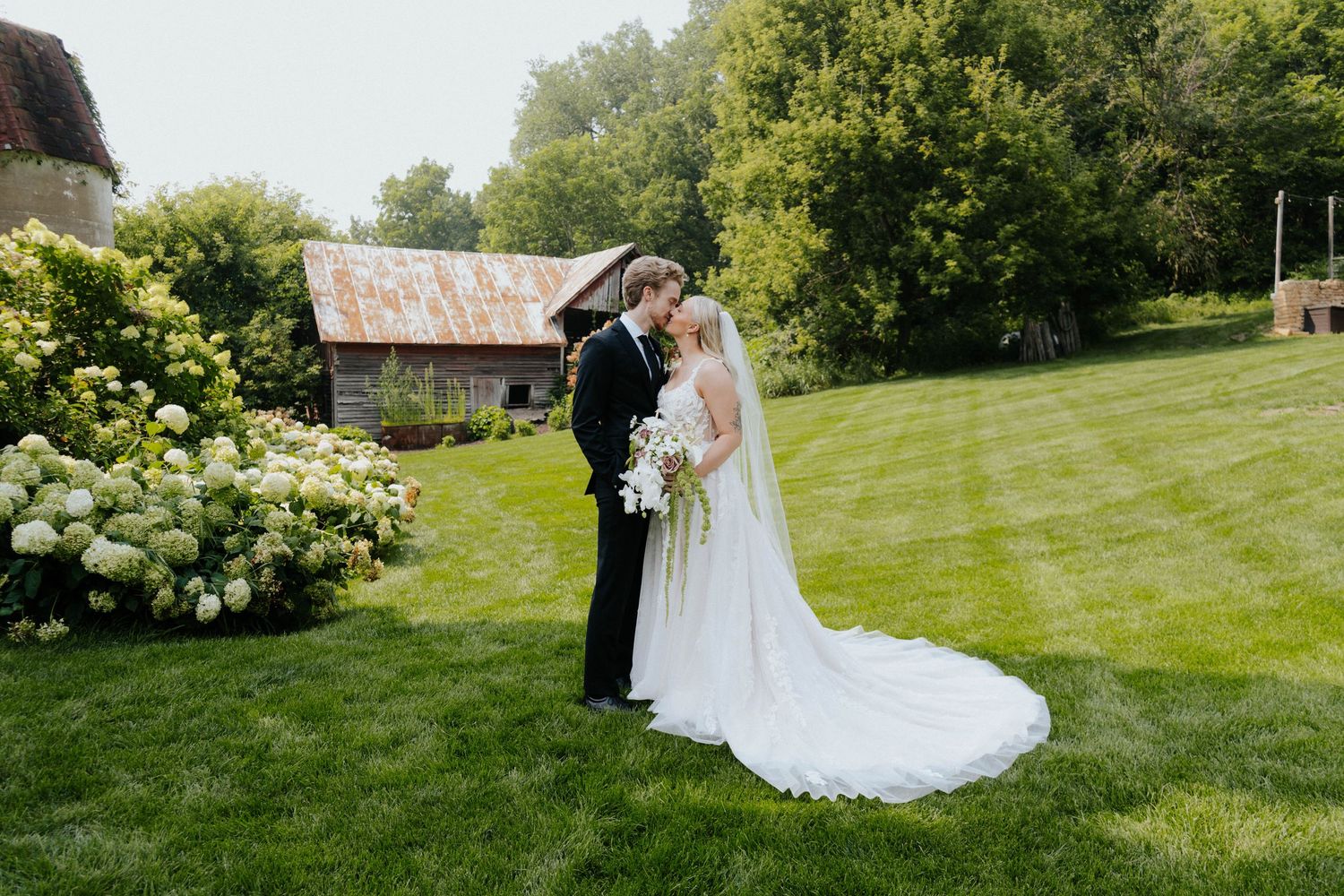 Bride and groom share a romantic kiss on a lush green lawn with a rustic barn and hydrangeas in the background.