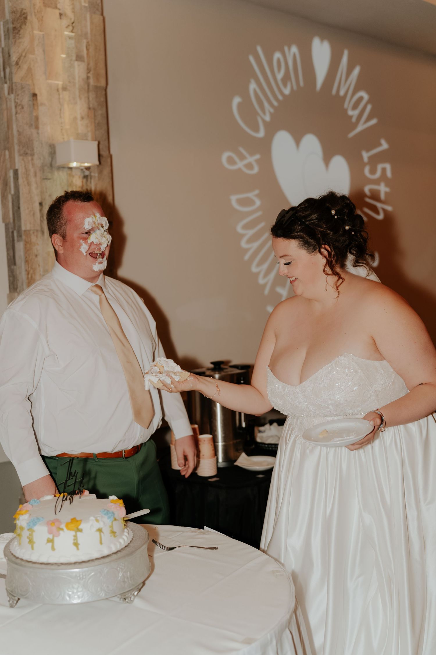 Bride smashes wedding cake into grooms face at reception, couple laughing, names projected on wall behind them.