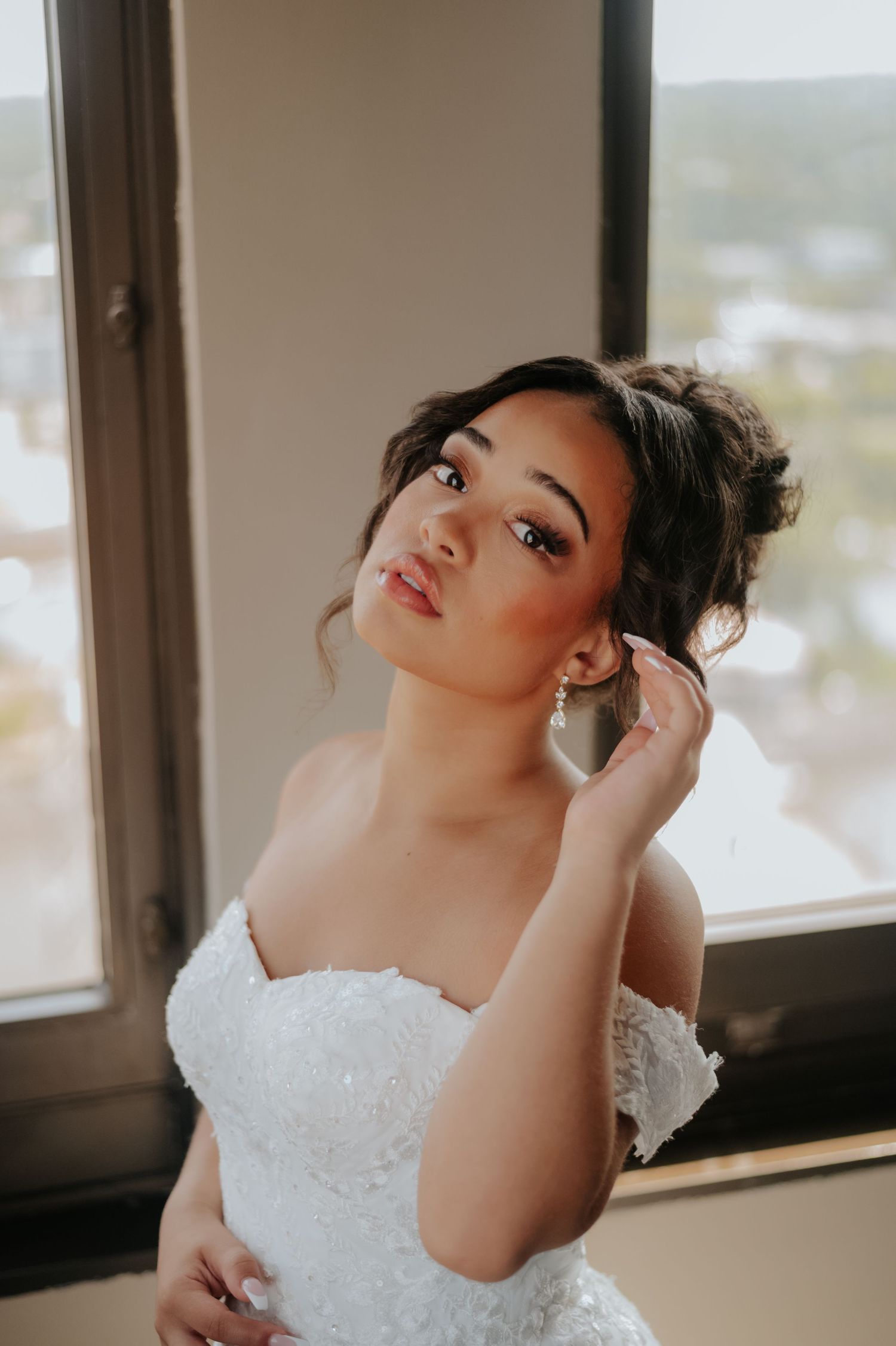 Bride in white lace off-shoulder gown adjusting pearl earring by window, elegant updo hairstyle and natural bridal makeup.