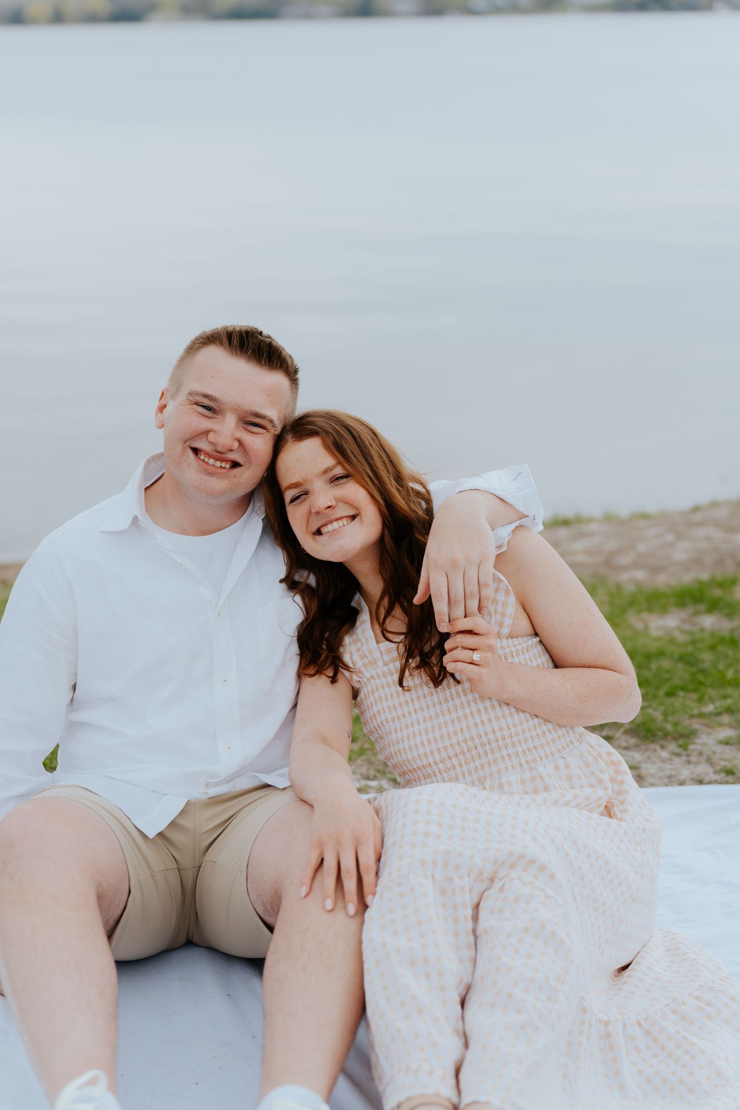 Smiling couple sitting on a blanket by the water, woman in floral dress and man in white shirt and khaki shorts.