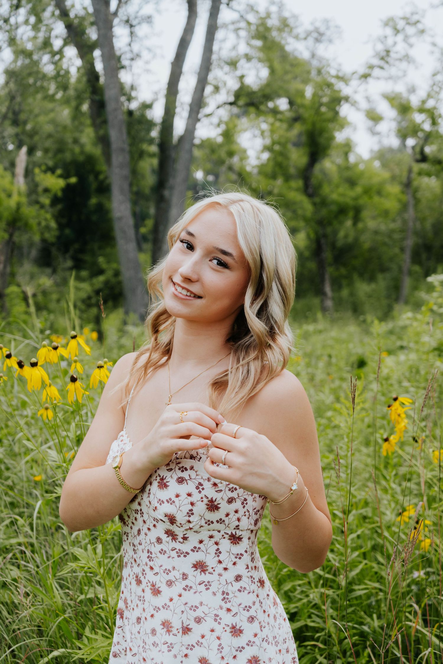 Smiling blonde woman in floral dress posing among wildflowers and yellow blooms in a green meadow.