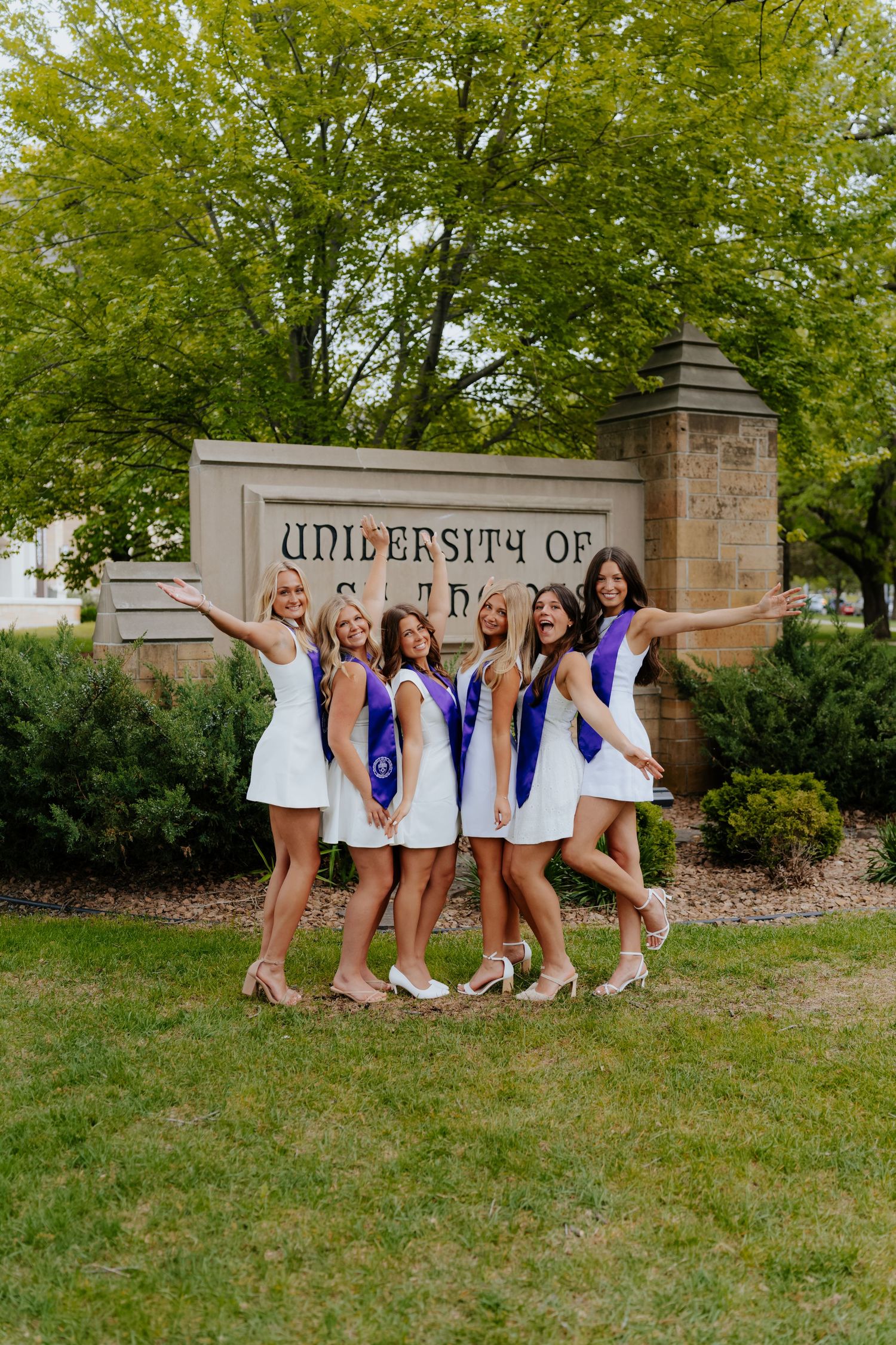 Six female graduates in white dresses and purple stoles pose joyfully in front of a University sign outdoors.