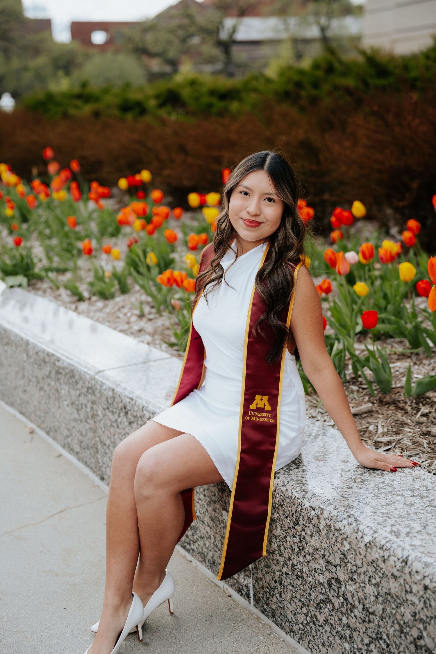 University of Minnesota graduate in white dress and maroon stole poses by colorful tulips outdoors.