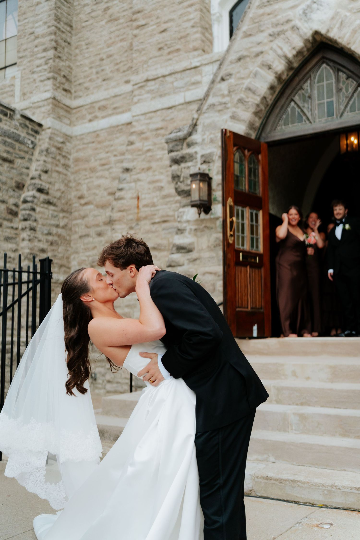 Groom dips bride for a kiss outside a stone church as wedding party cheers on the steps behind them.