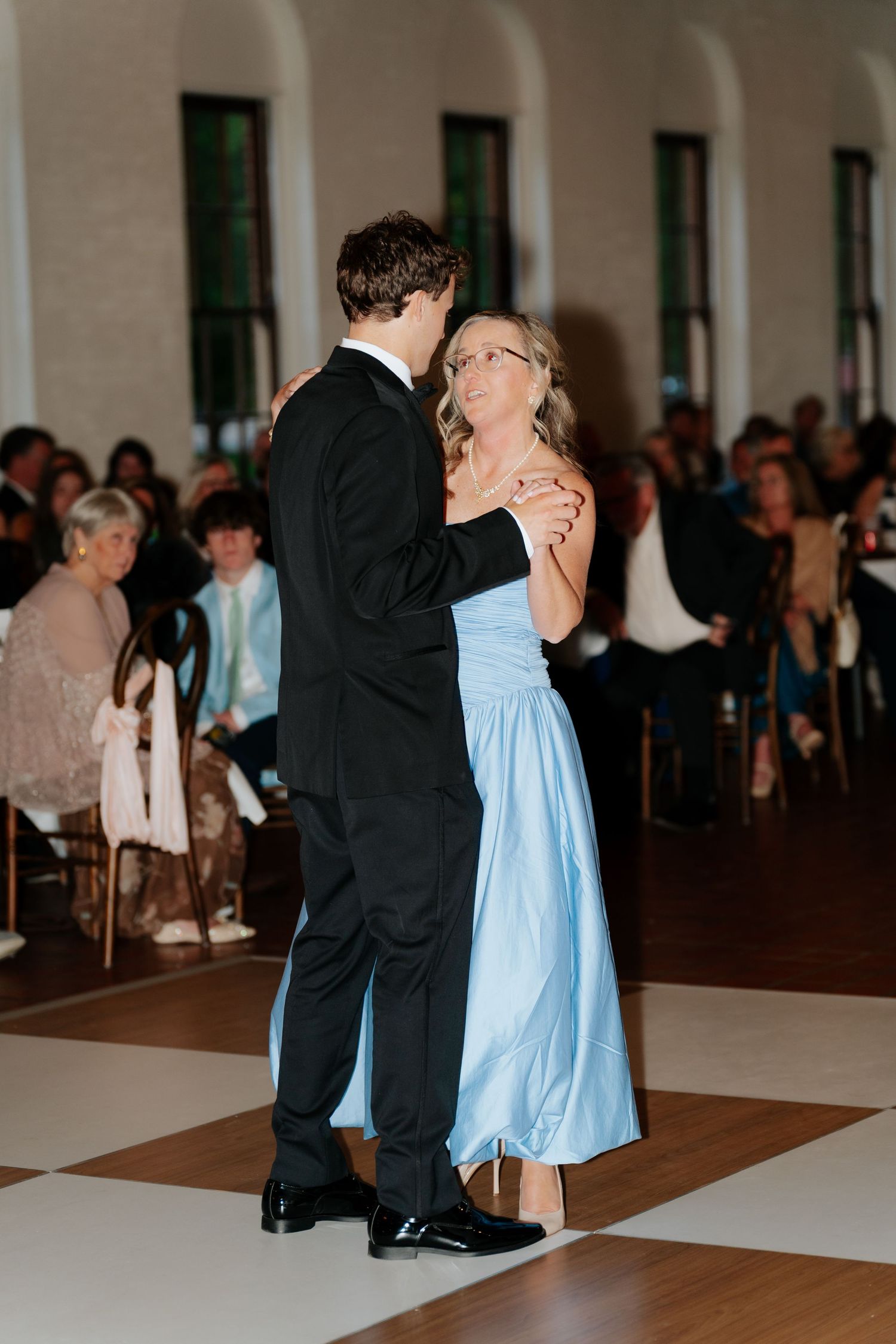 A groom in a black tuxedo shares a mother-son dance with a woman in a light blue gown at a wedding reception.