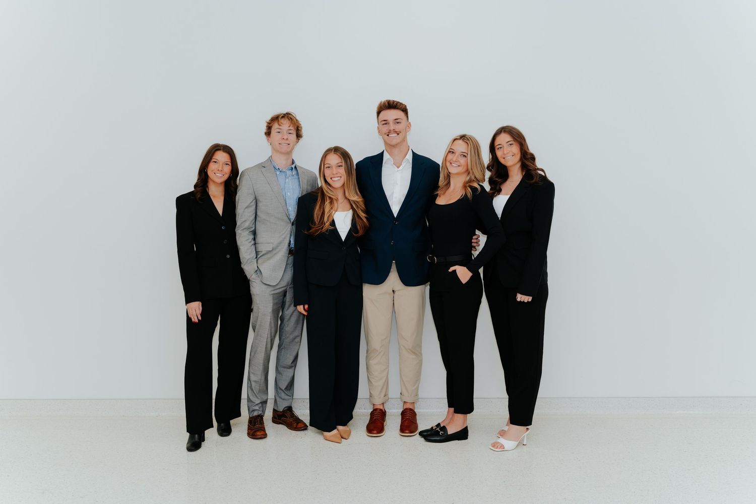 Six professionally dressed young adults smiling together in a group photo against a light gray background.