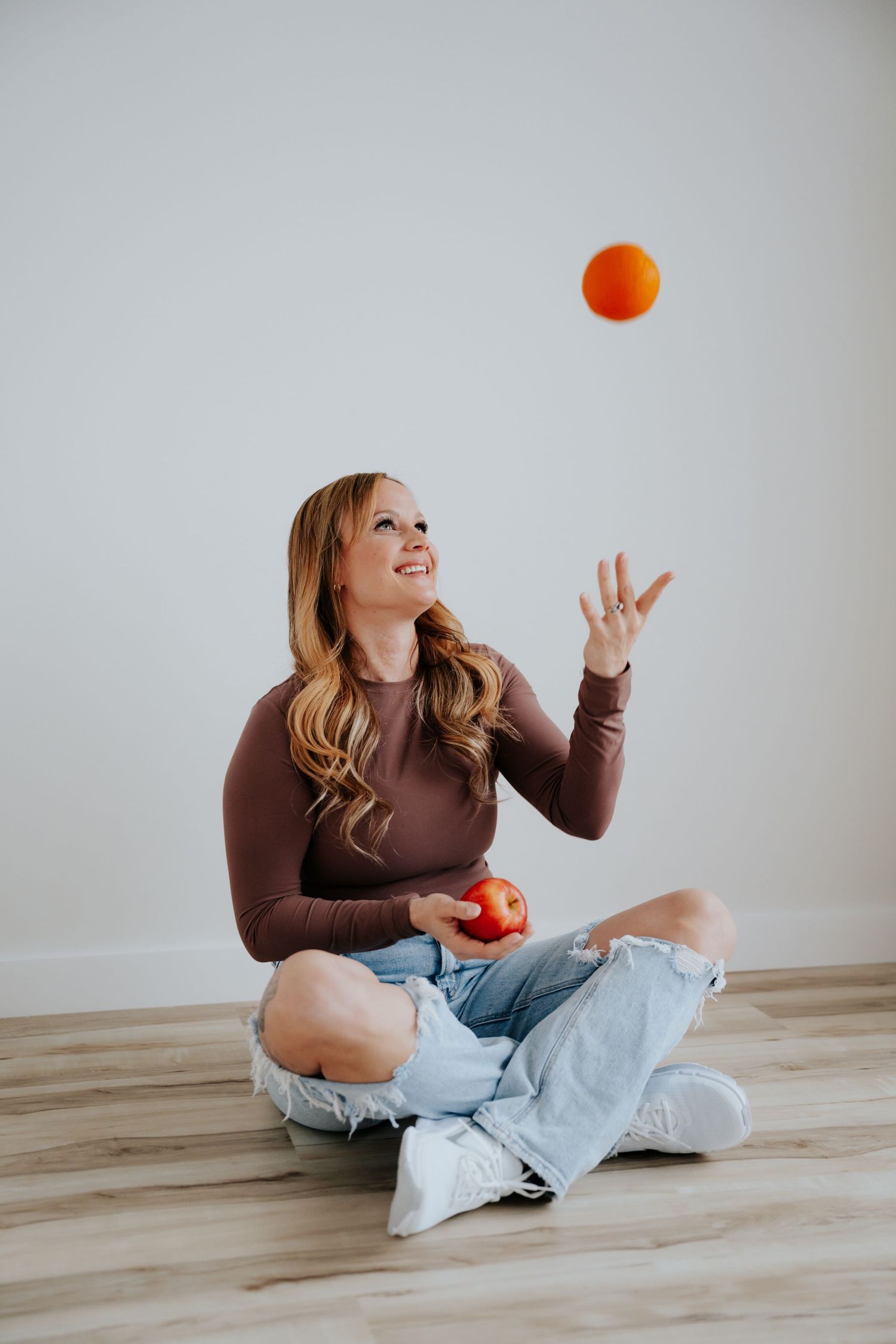 Woman sitting cross-legged on wood floor tossing an orange in the air while holding an apple, wearing jeans and brown top.