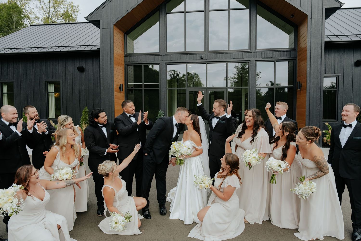 Wedding party celebrating outside modern black barn venue, bridesmaids in champagne gowns, groomsmen in black tuxedos.
