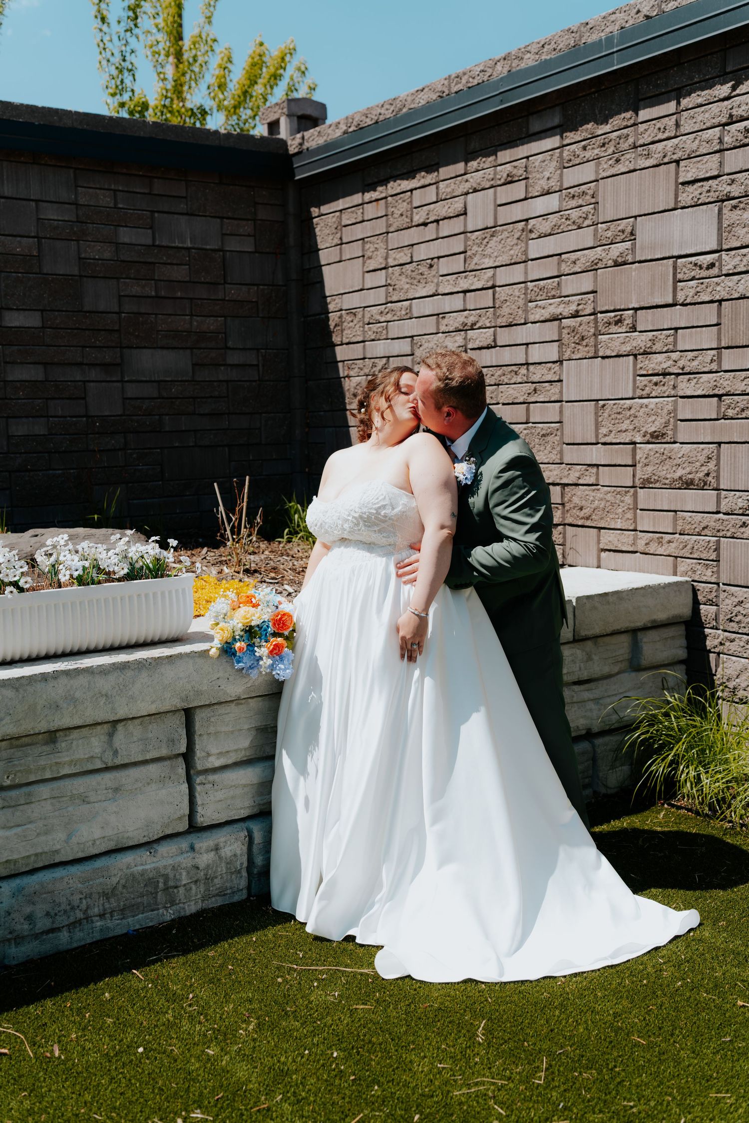 A bride and groom share a kiss outdoors in a garden setting, bride holding a colorful bouquet in a white gown.