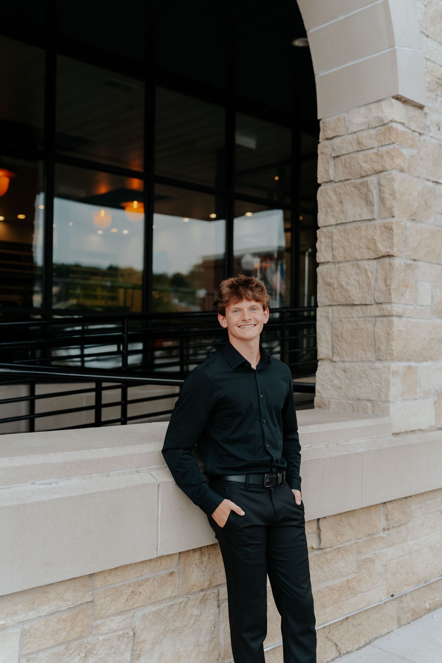 Young man in black outfit smiling confidently outside a modern stone building with large windows.